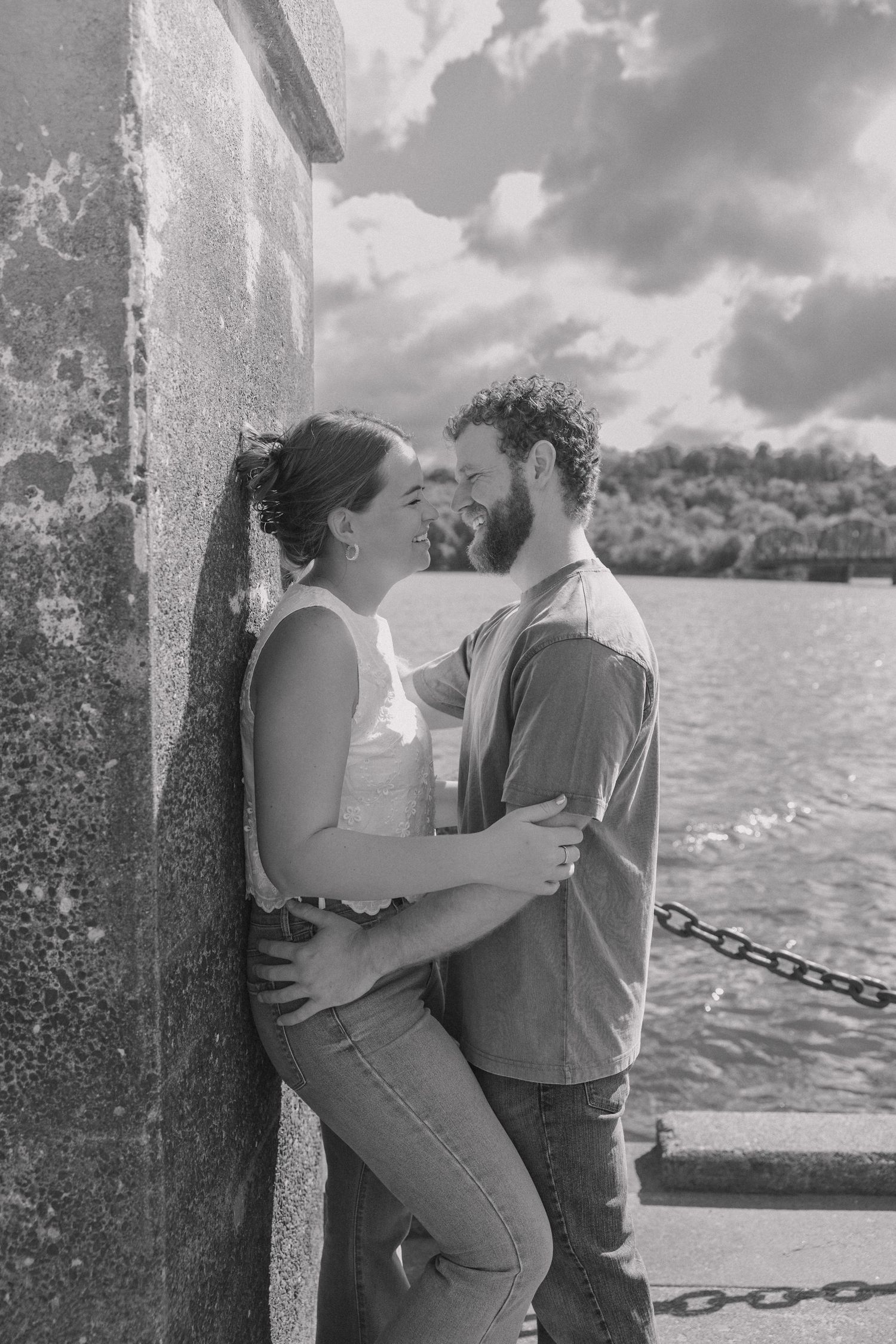 Black and white photo of a couple laughing and embracing against a stone wall near a waterfront on a cloudy day.