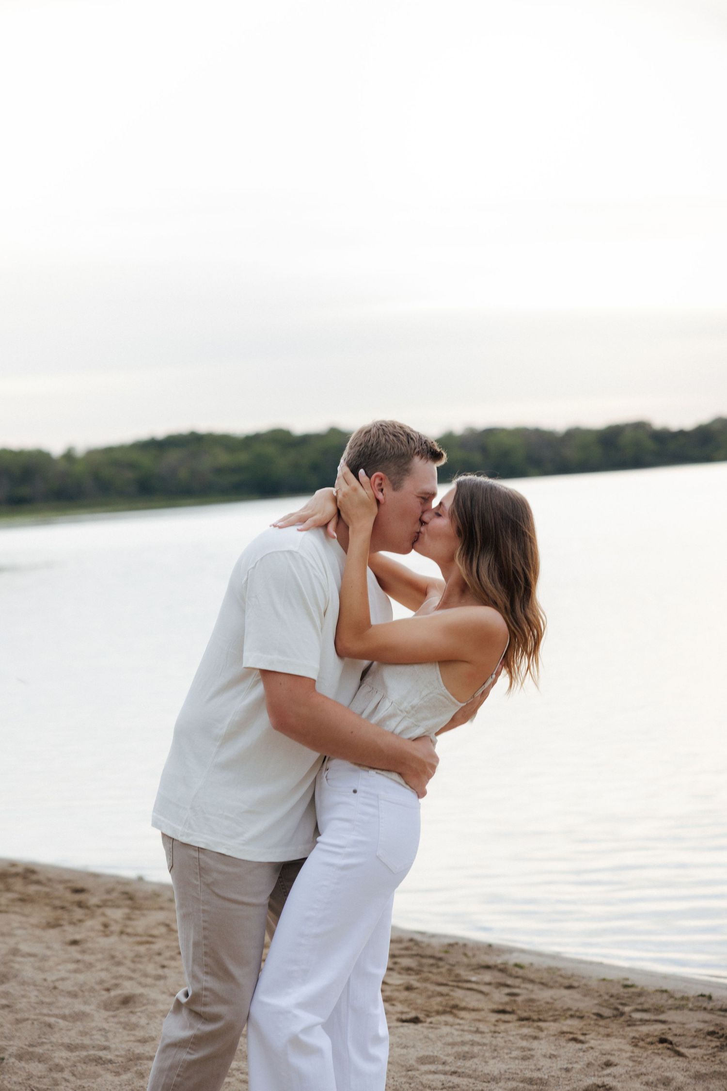 Couple sharing a romantic kiss on a sandy beach by a calm lake, dressed in white and beige outfits.