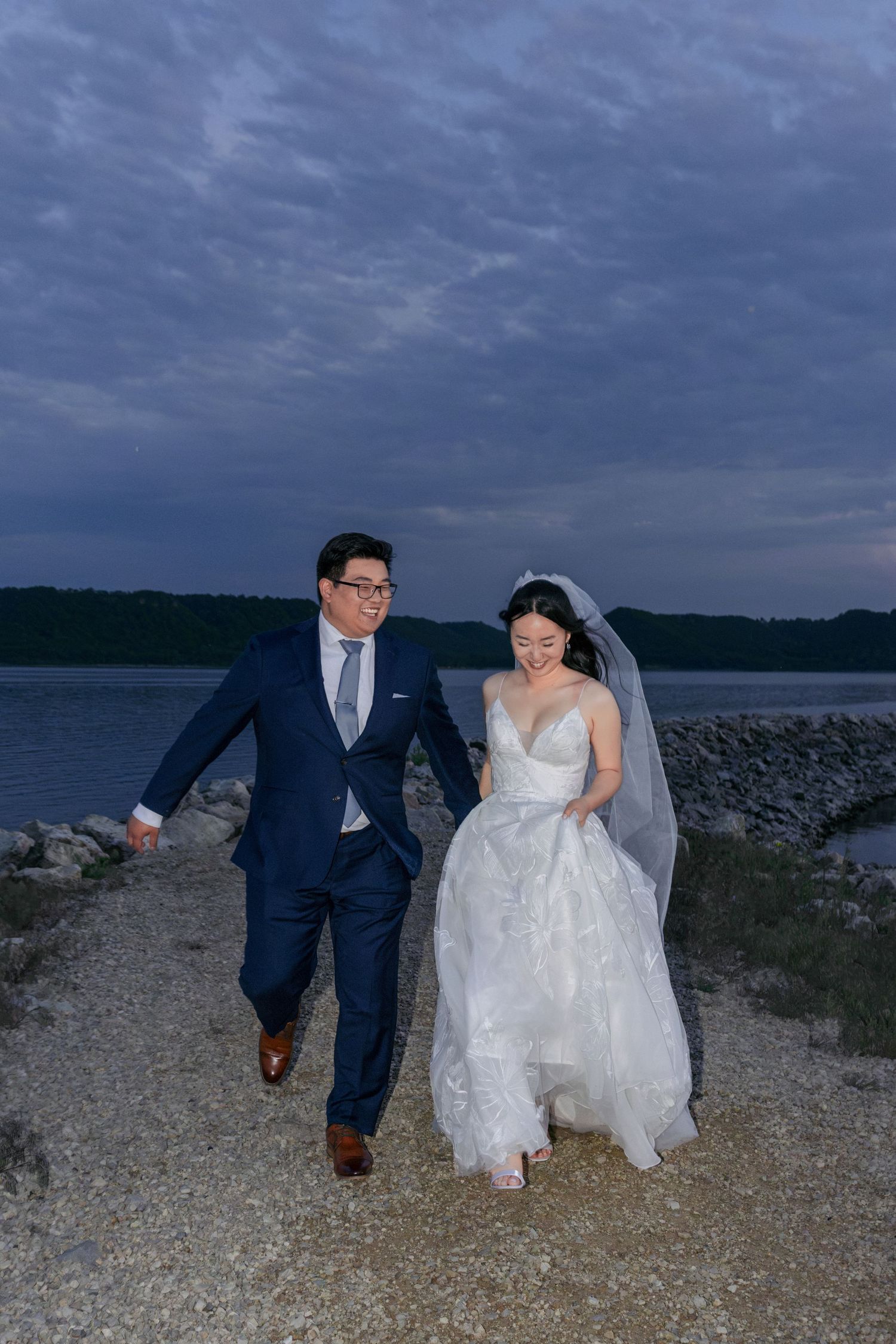 Bride and groom walk hand in hand along a rocky waterfront path under a dramatic evening sky.