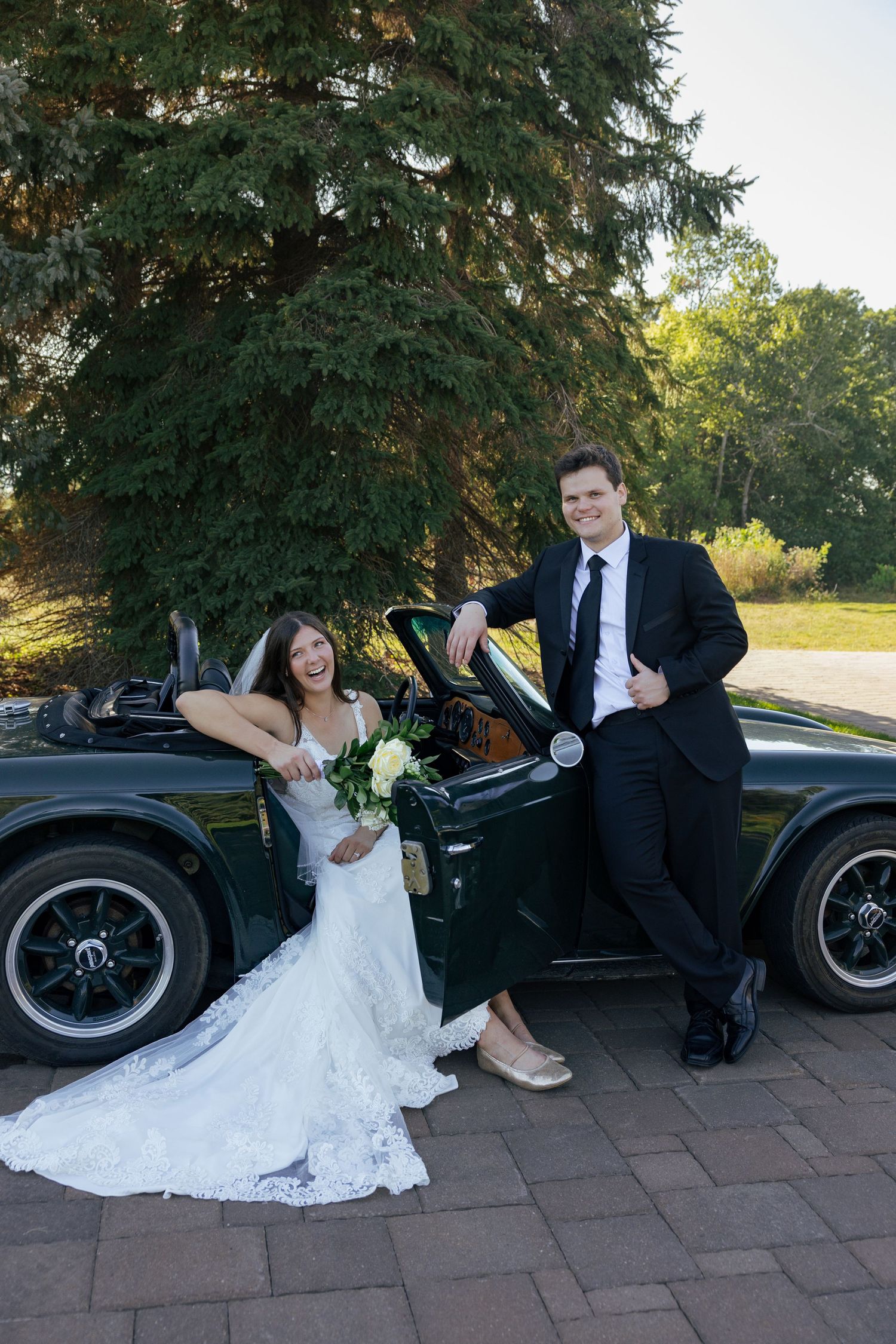 Laughing bride and groom posing beside classic green convertible car outdoors.