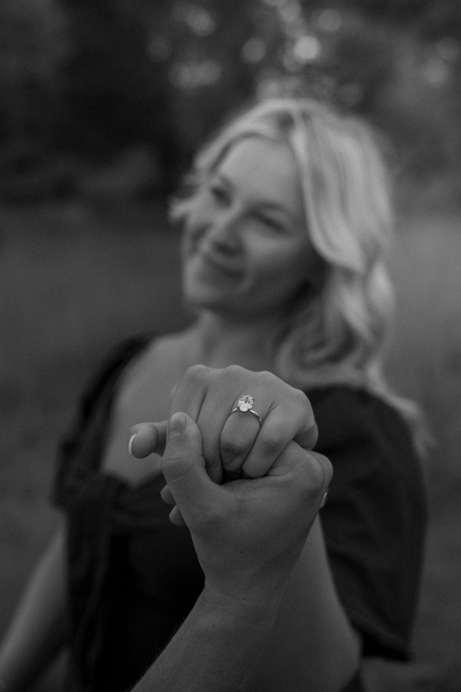 Black and white photo of a smiling blonde woman showing off her engagement ring outdoors.