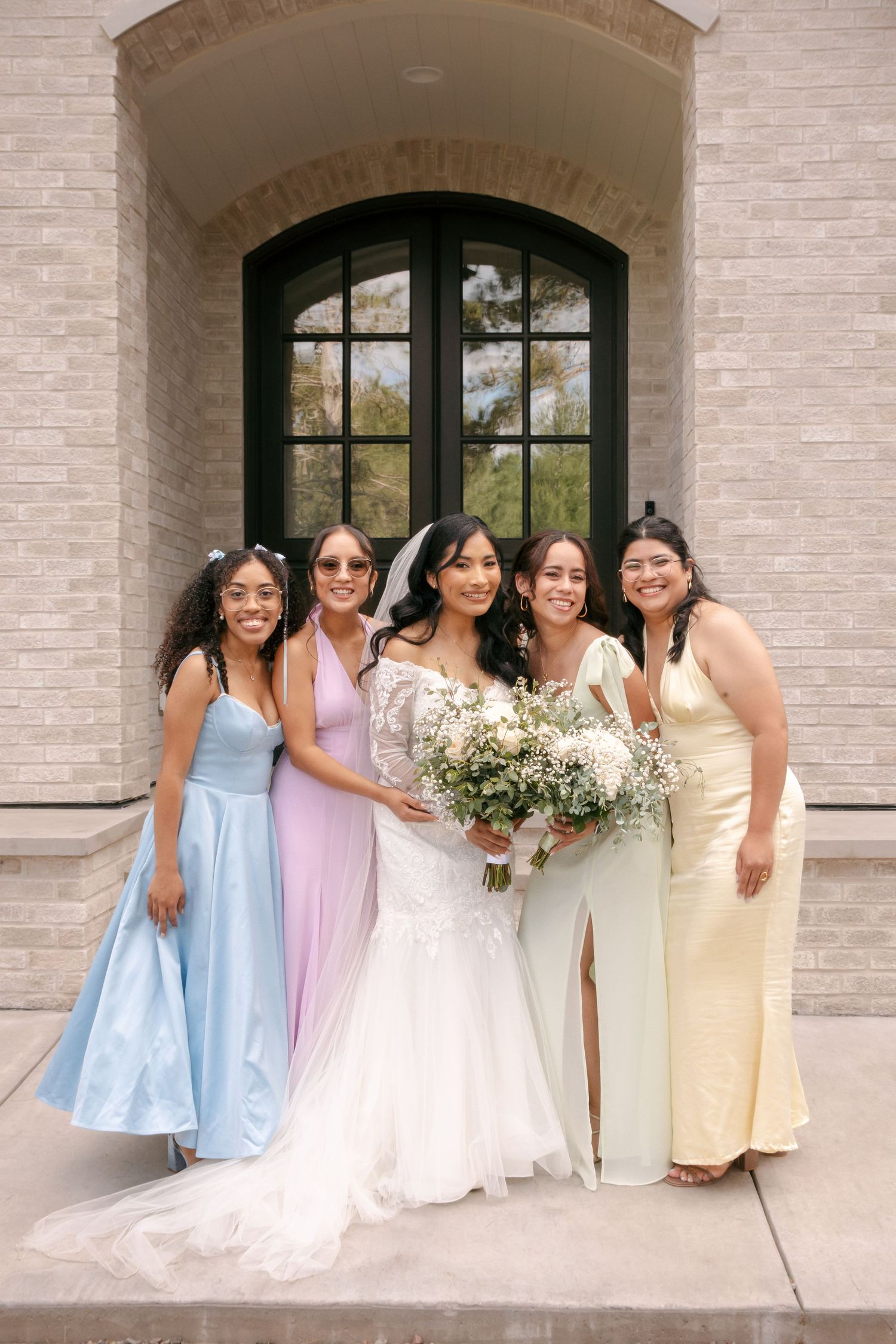 Bride in lace gown poses with four bridesmaids in colorful pastel dresses holding a white bouquet.