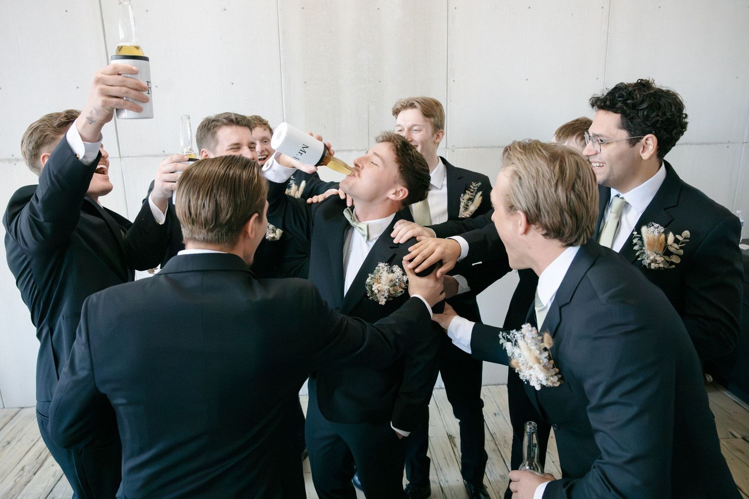 Groomsmen in black suits celebrating with drinks, toasting and laughing together at a wedding reception.