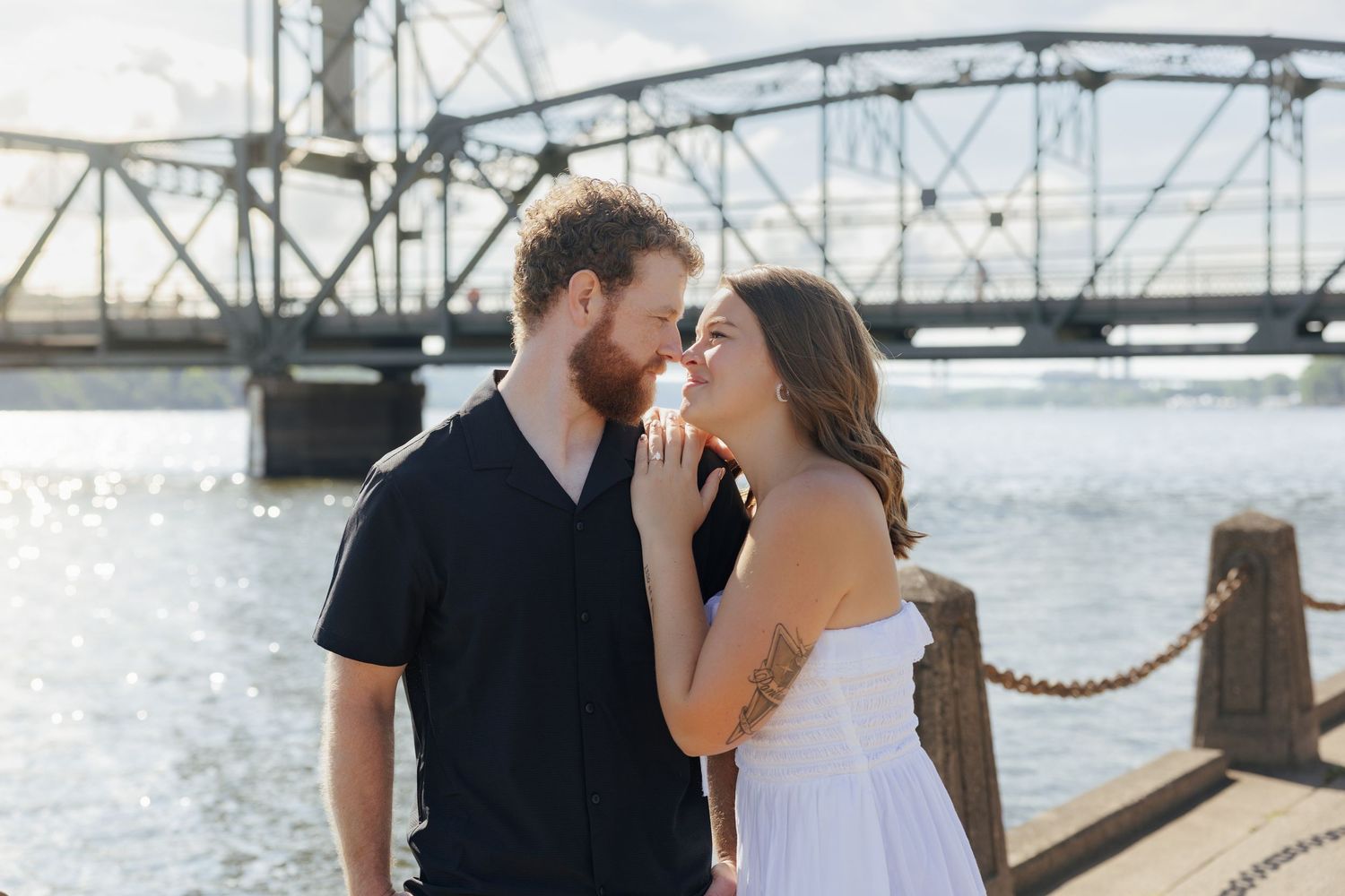 Couple sharing a romantic moment on a waterfront dock with a steel bridge and sunlit river in the background.
