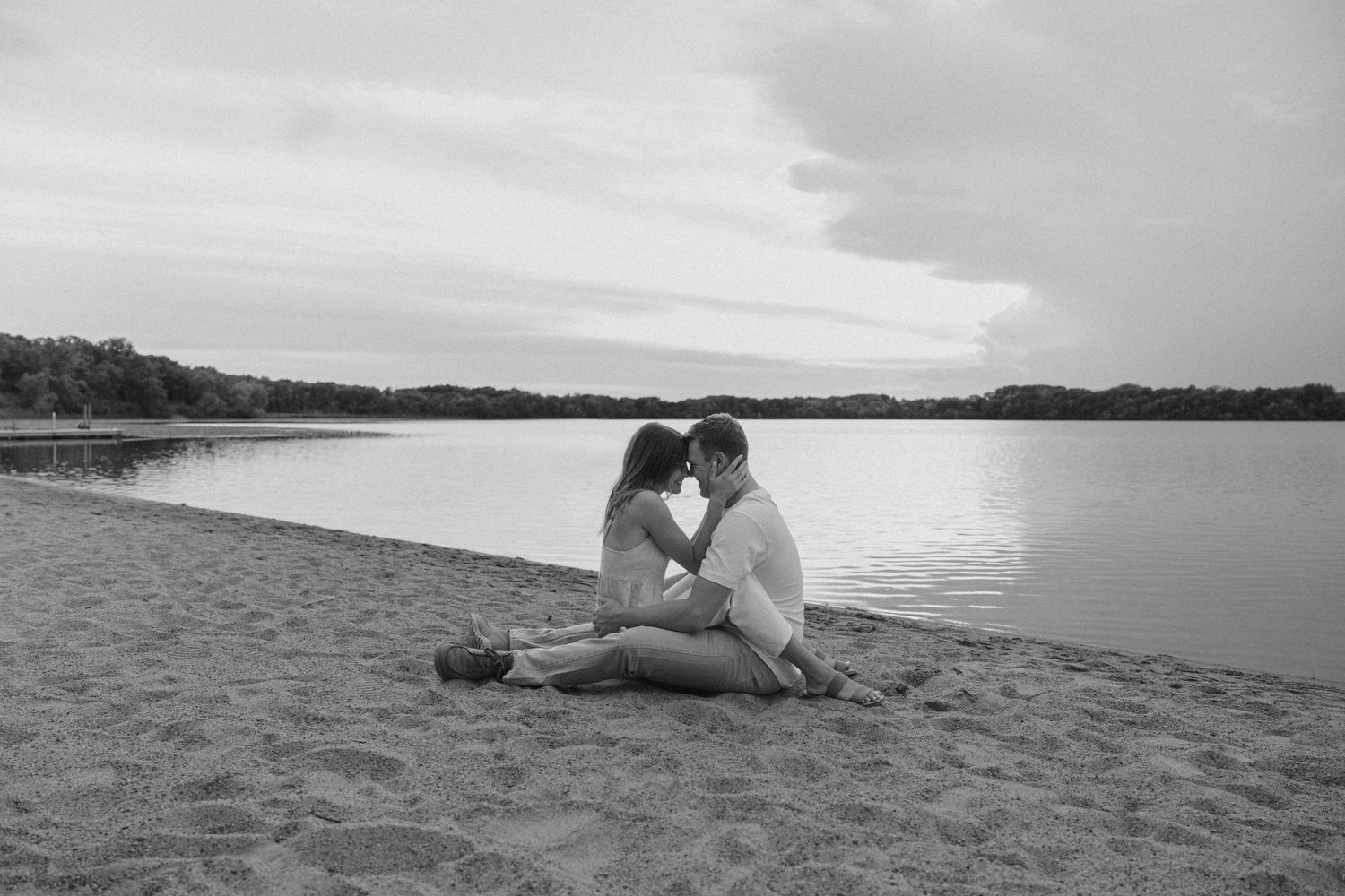 Black and white photo of a romantic couple sitting close together on a sandy beach by a calm lake at sunset.