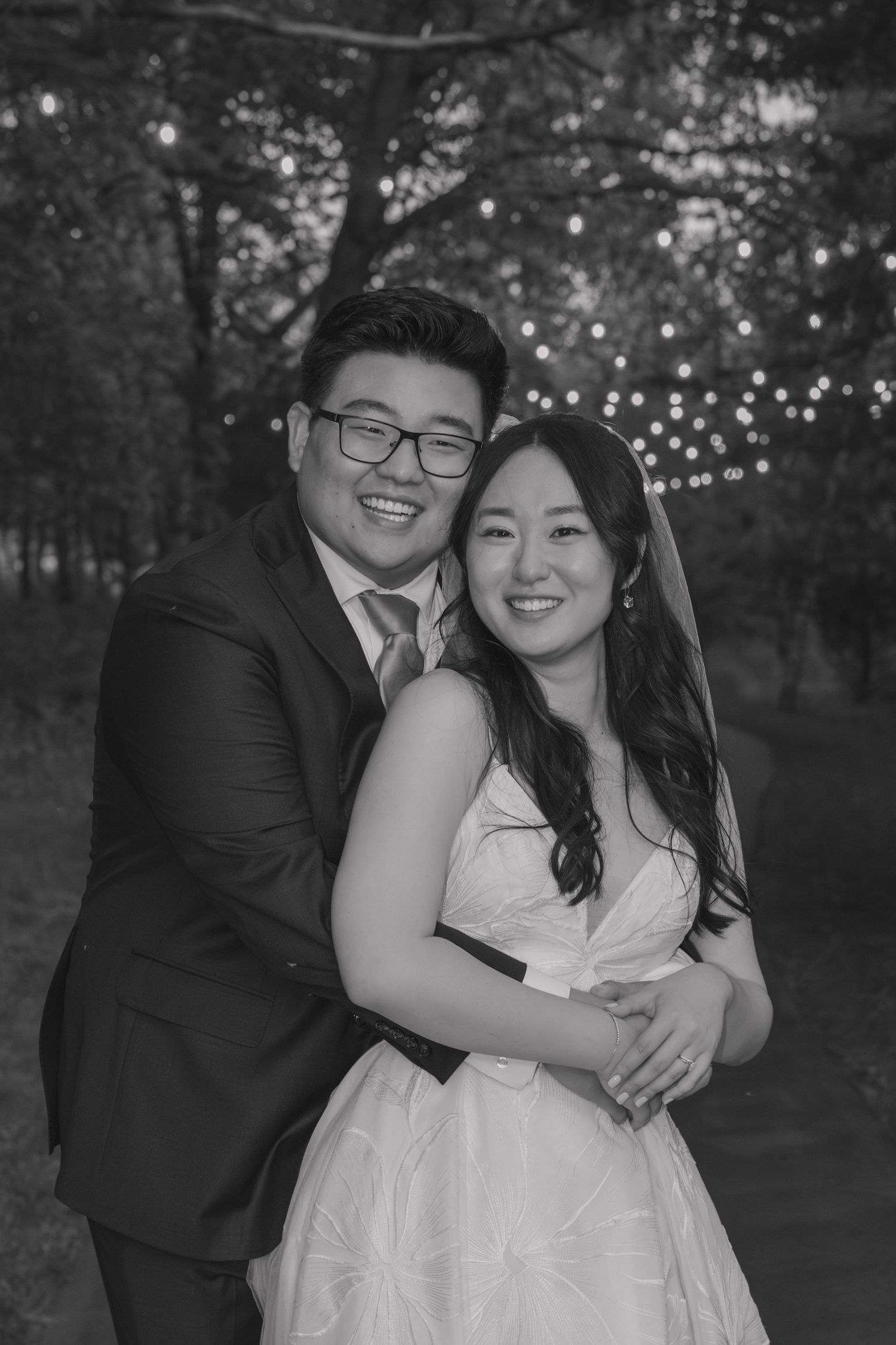 Black and white portrait of smiling bride and groom embracing outdoors with twinkling string lights behind them.