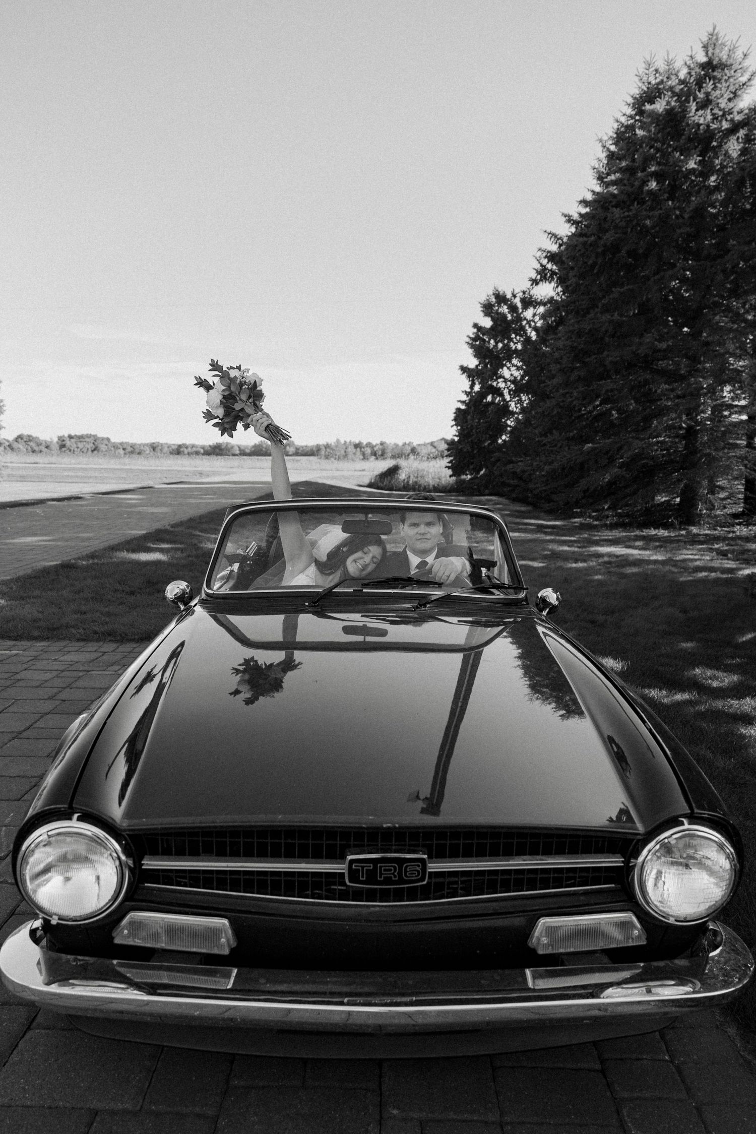 Black and white photo of a bride and groom in a vintage convertible car, bride holding bouquet aloft.