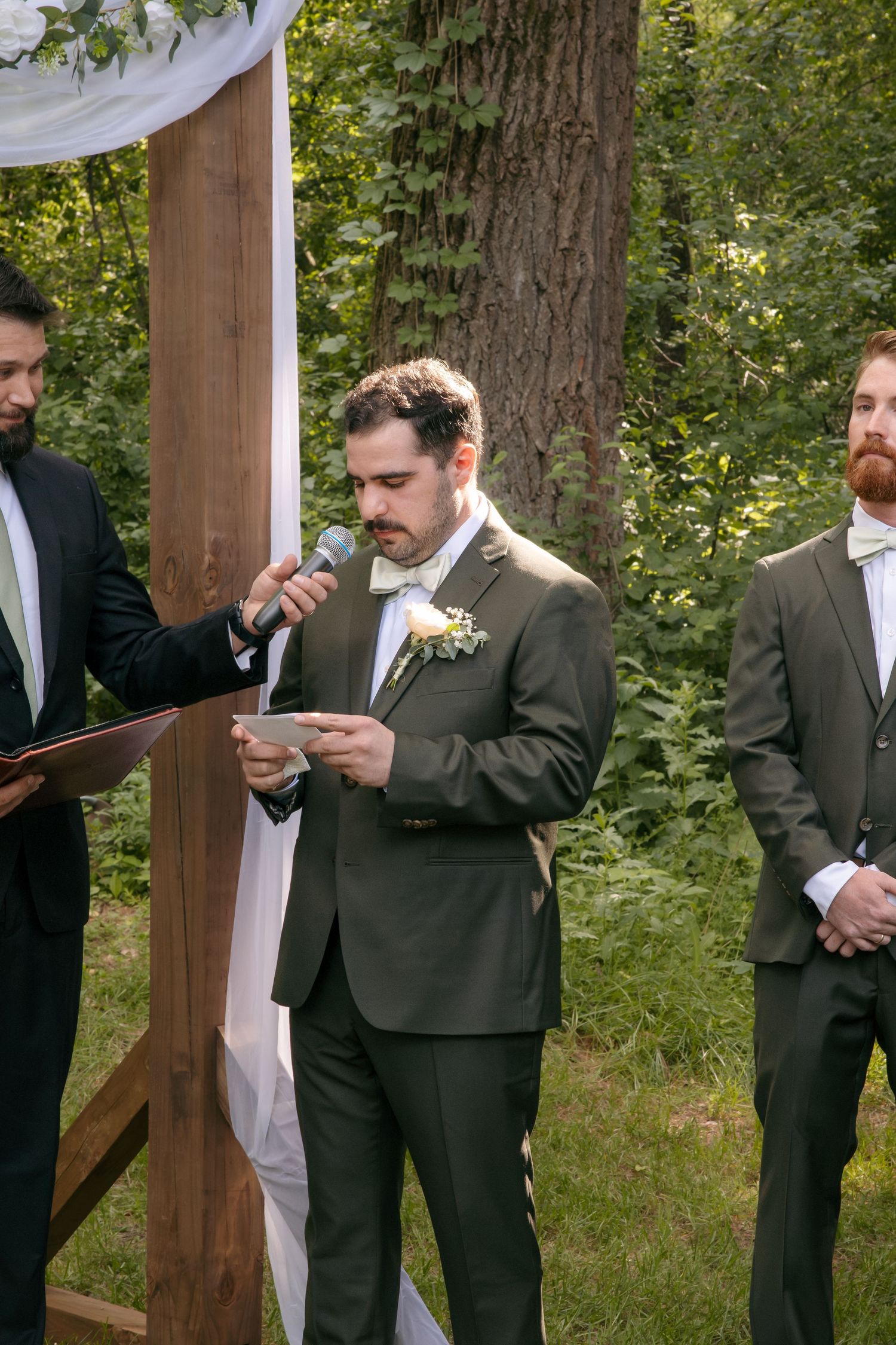 Groom reads wedding vows from a card at an outdoor ceremony beside a wooden arch with white draping.