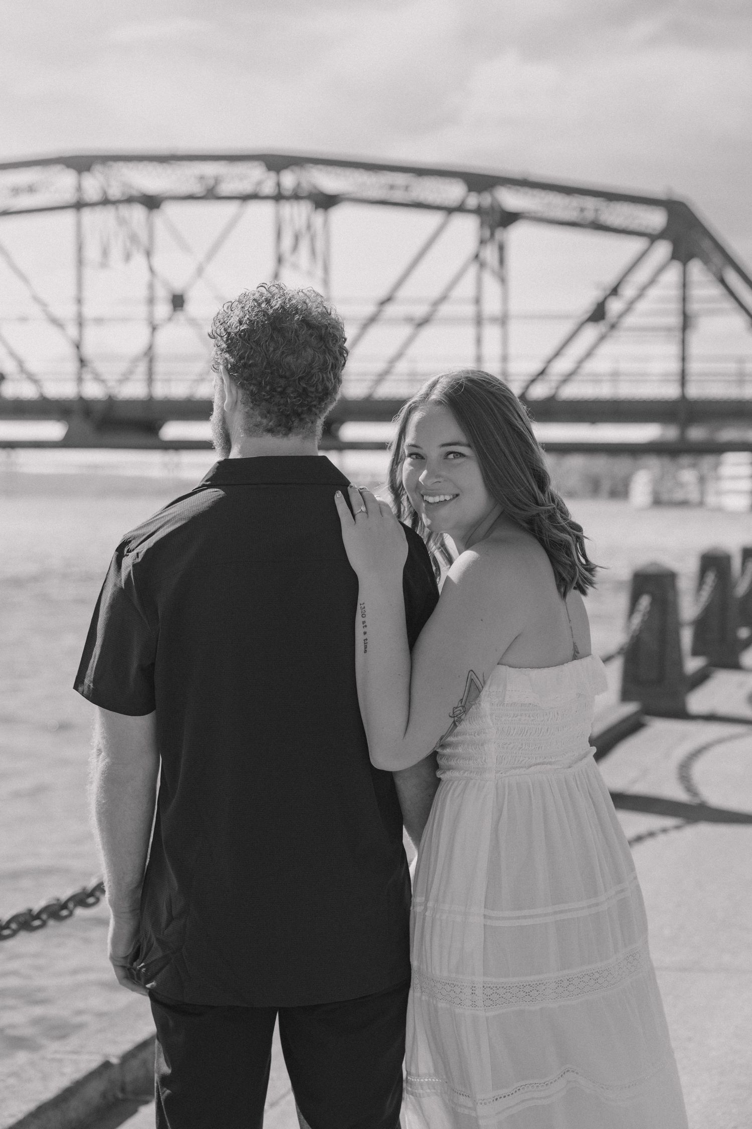 Black and white photo of a smiling woman in a white dress with a man near a bridge waterfront.