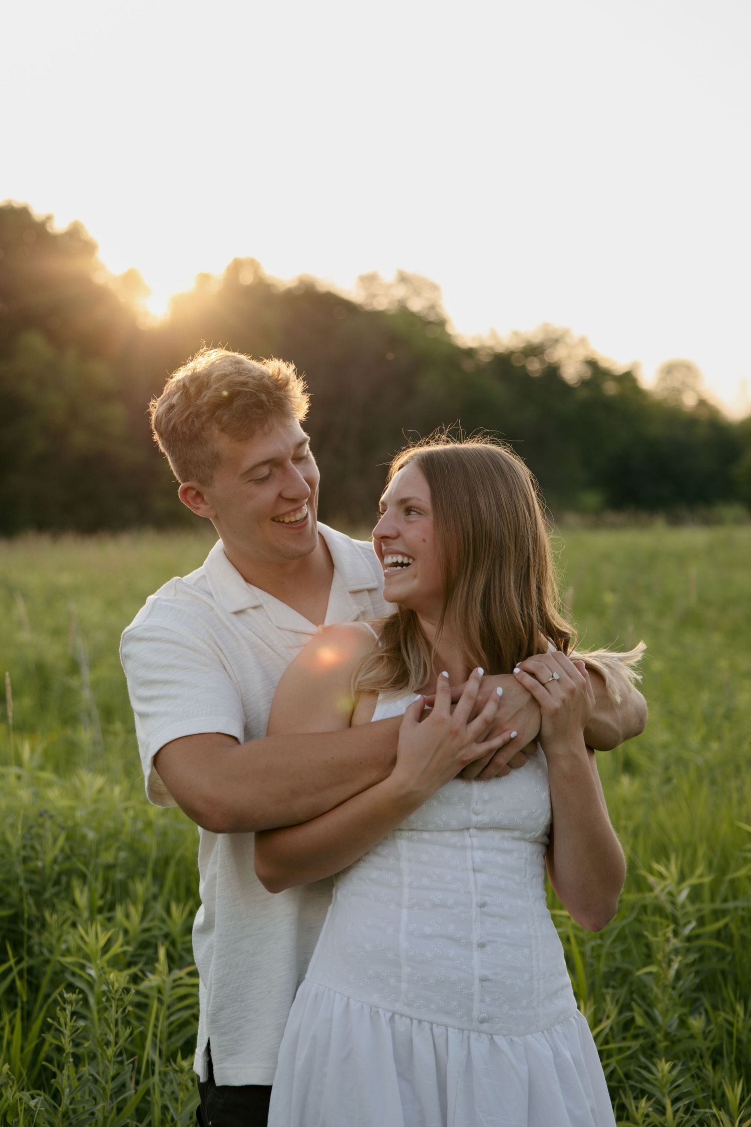A happy couple embraces in a sunlit green field at golden hour, both wearing white outfits and smiling joyfully.