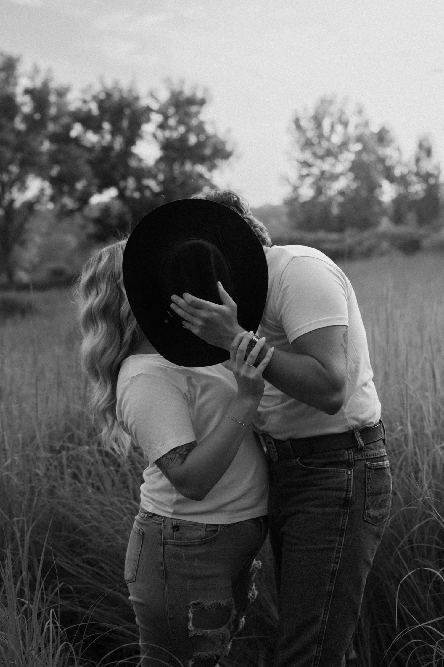 Black and white photo of a couple kissing behind a cowboy hat in a field of tall grass.