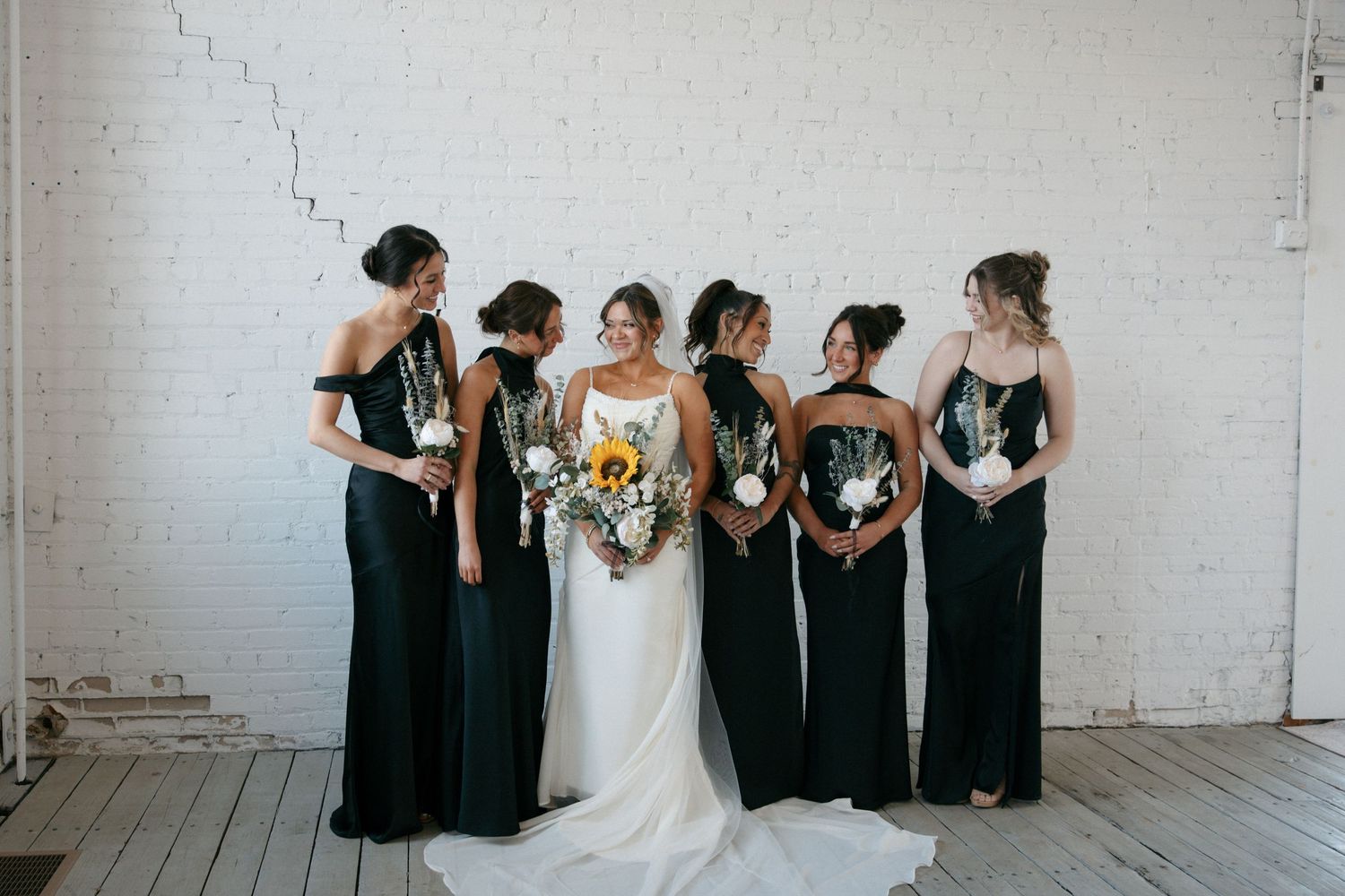 Bride in white gown with bridesmaids in dark green dresses holding bouquets against a white brick wall.
