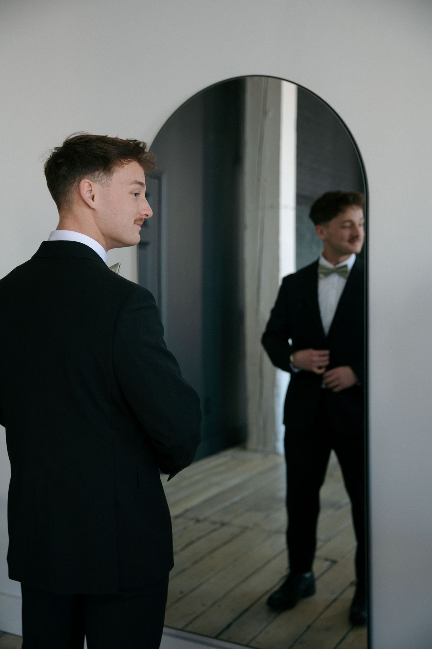 Dapper groom in a black tuxedo admiring his reflection in an arched full-length mirror before the wedding.