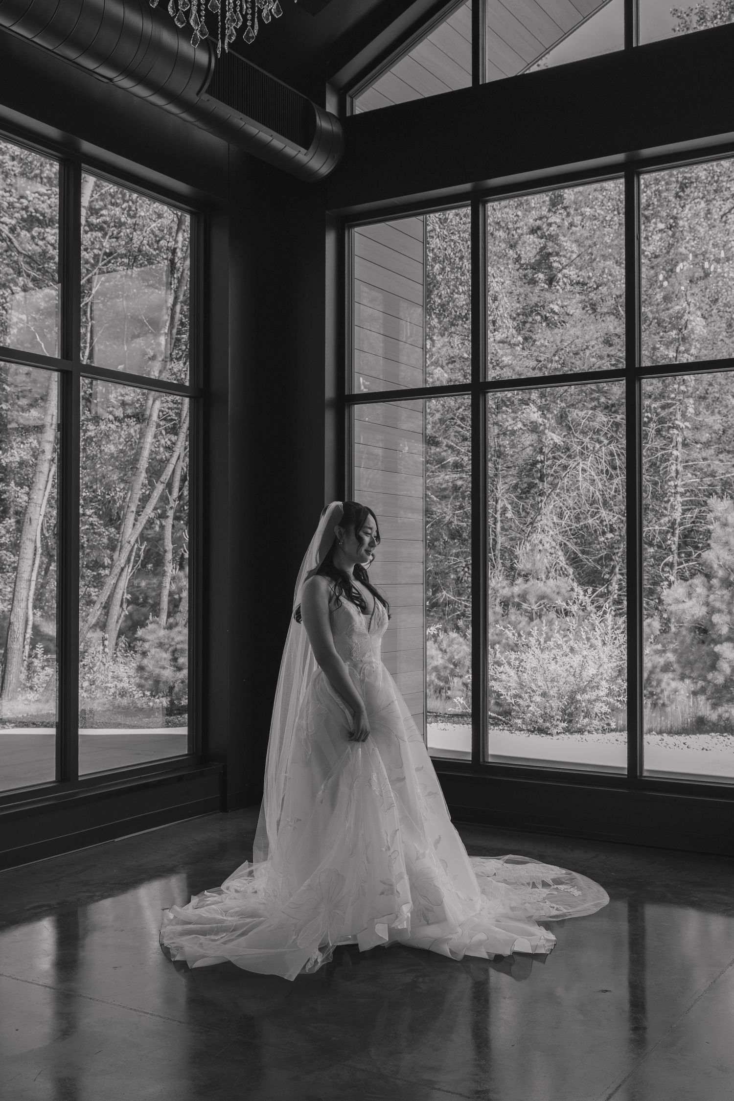 Bride in a flowing white gown and veil stands before large windows with lush forest views in black and white.