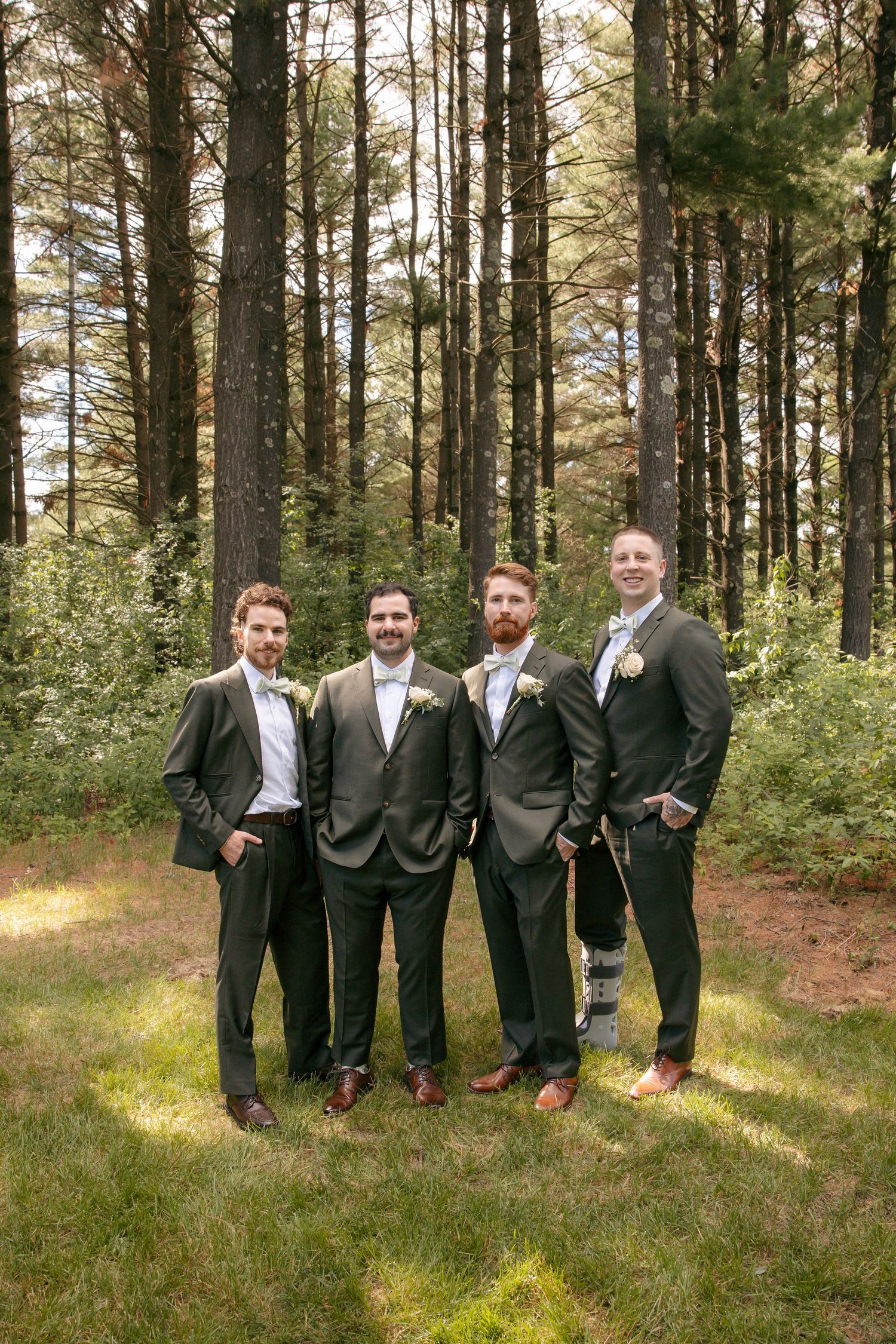 Four groomsmen in dark suits with boutonnieres pose outdoors in a pine forest setting for a wedding party photo.