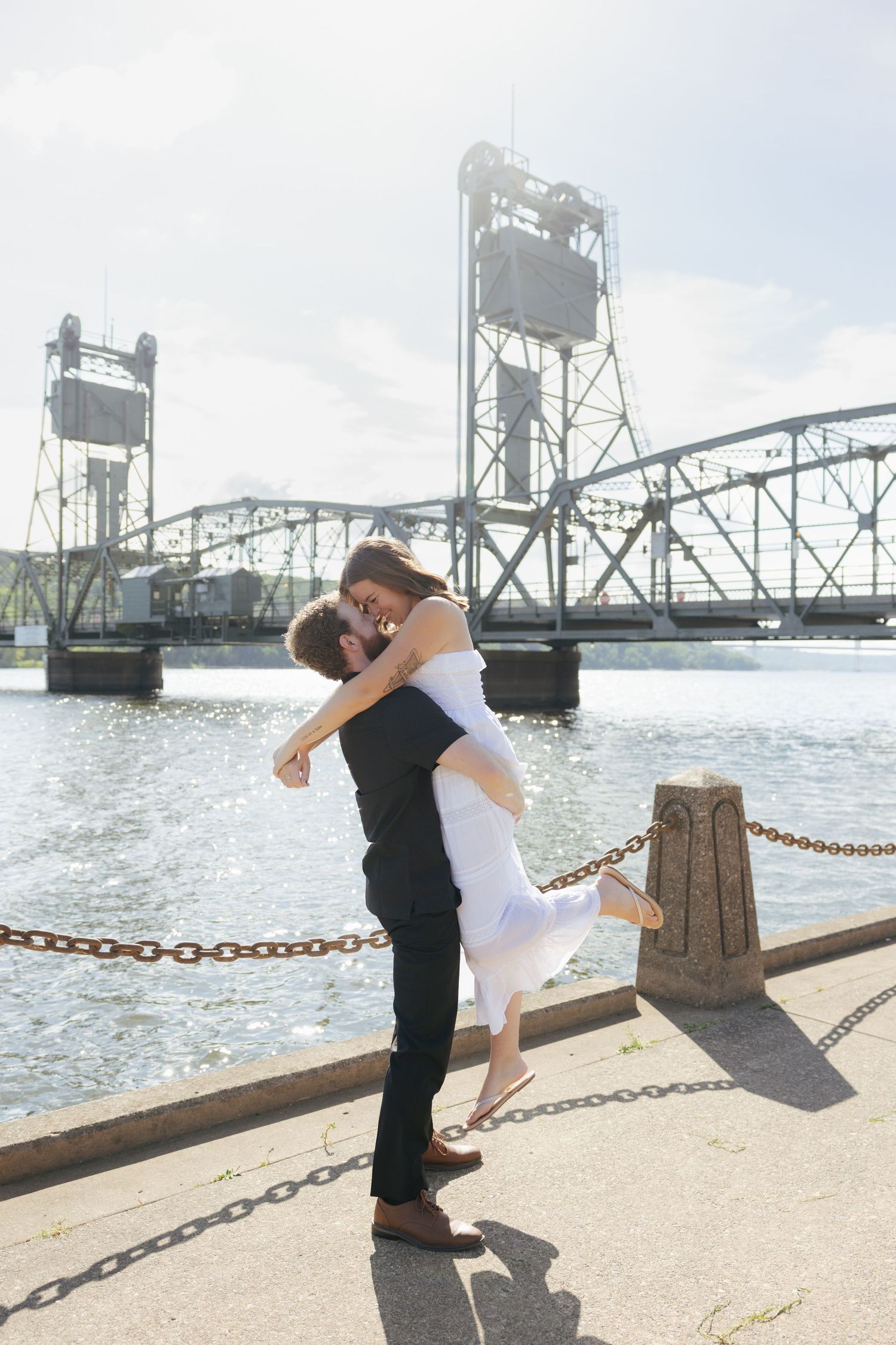 Couple sharing a romantic kiss as man lifts woman in white dress by a waterfront bridge on a sunny day.