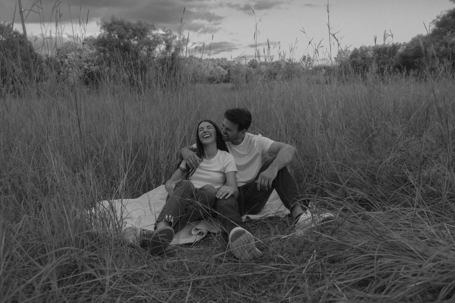 Couple sitting together in tall grass field, sharing a joyful moment on a blanket in black and white photo.