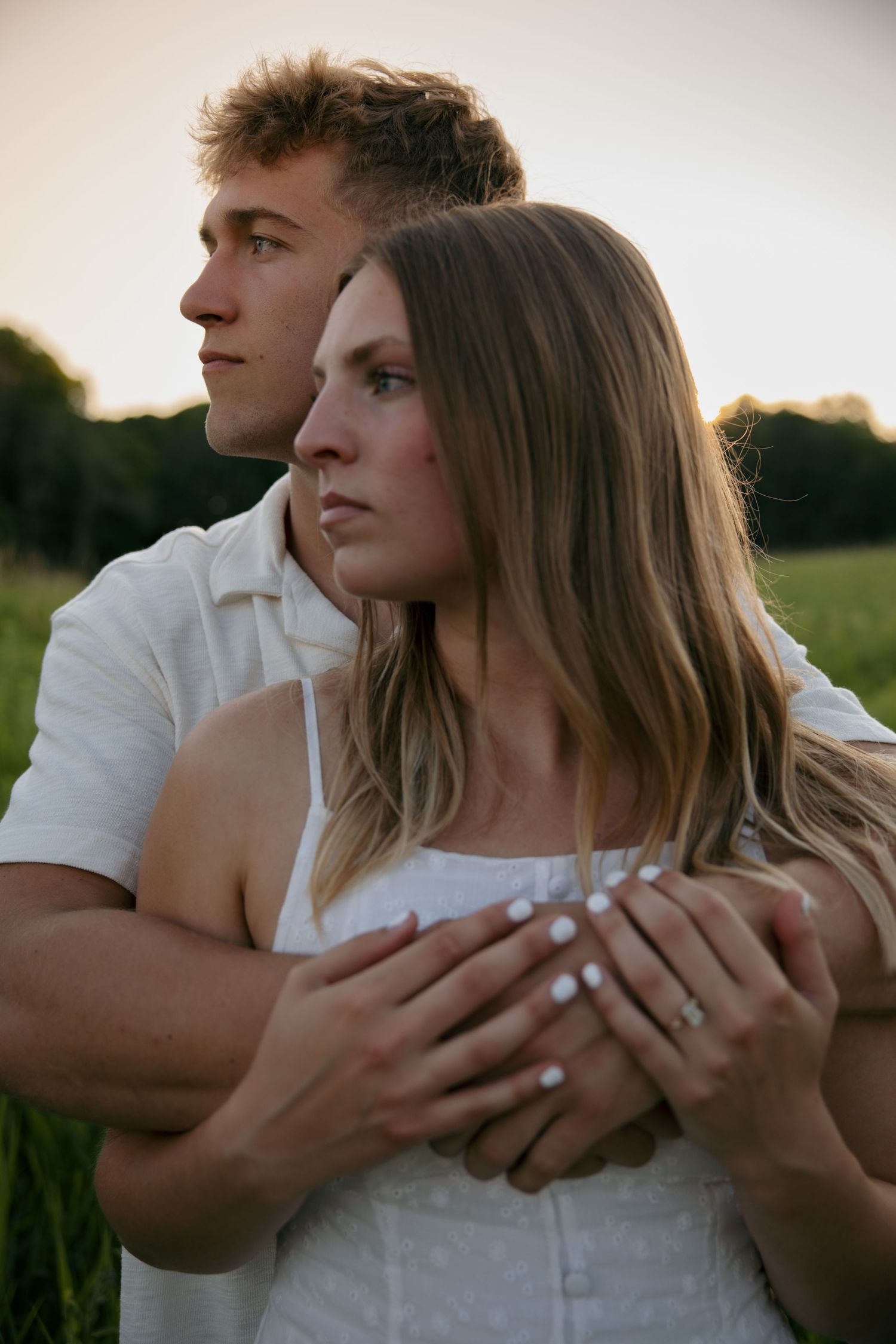 Young couple embracing in a green field at sunset, man in white shirt holding woman in white dress from behind.