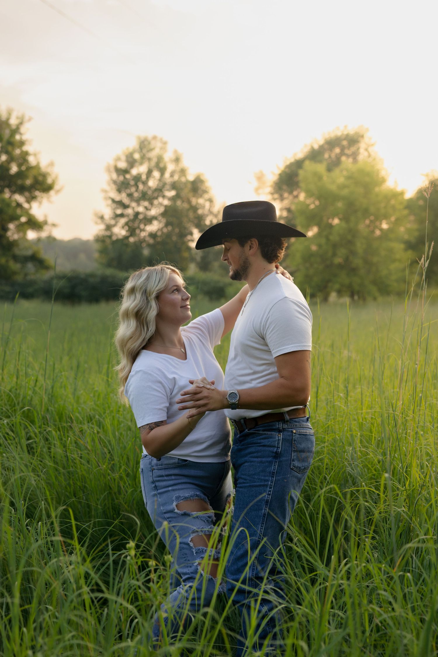 Couple embracing in lush green field at golden hour, woman in ripped jeans, man in cowboy hat and white tee.