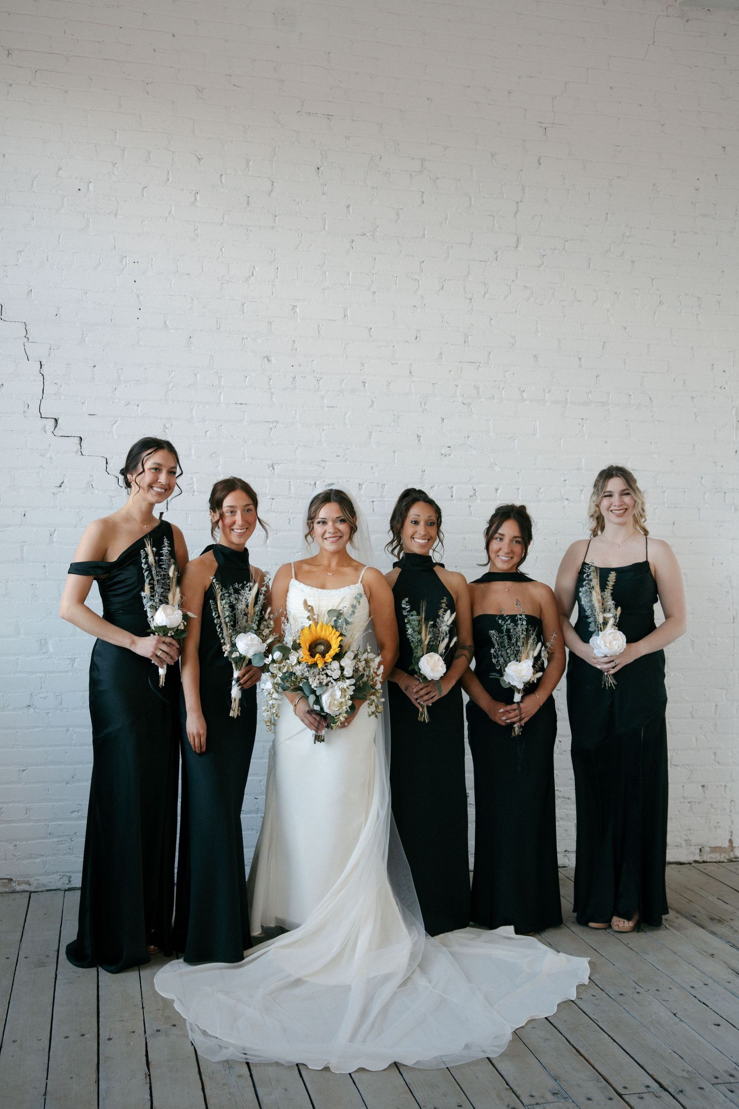 Bride in white gown with five bridesmaids in black dresses holding bouquets against white brick wall.