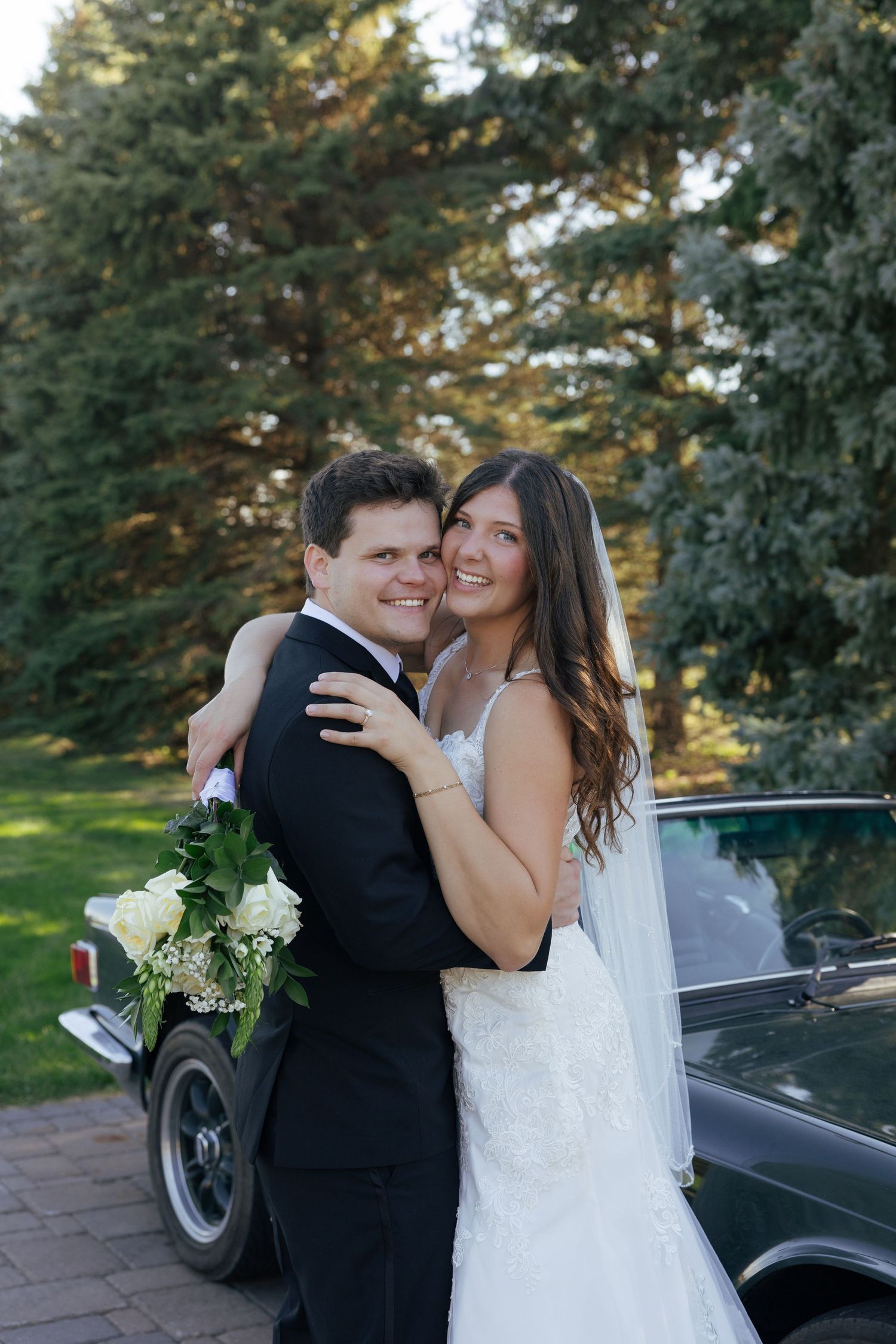 Bride and groom embrace smiling in wedding attire beside a vintage car with lush green trees in background.