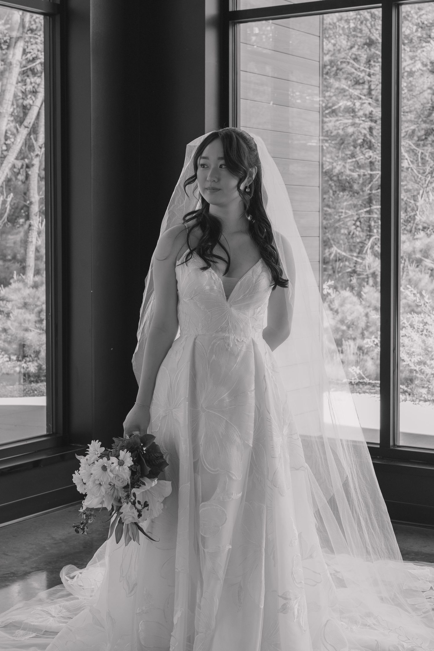 Black and white portrait of a bride in a lace gown and veil holding a bouquet by large windows.