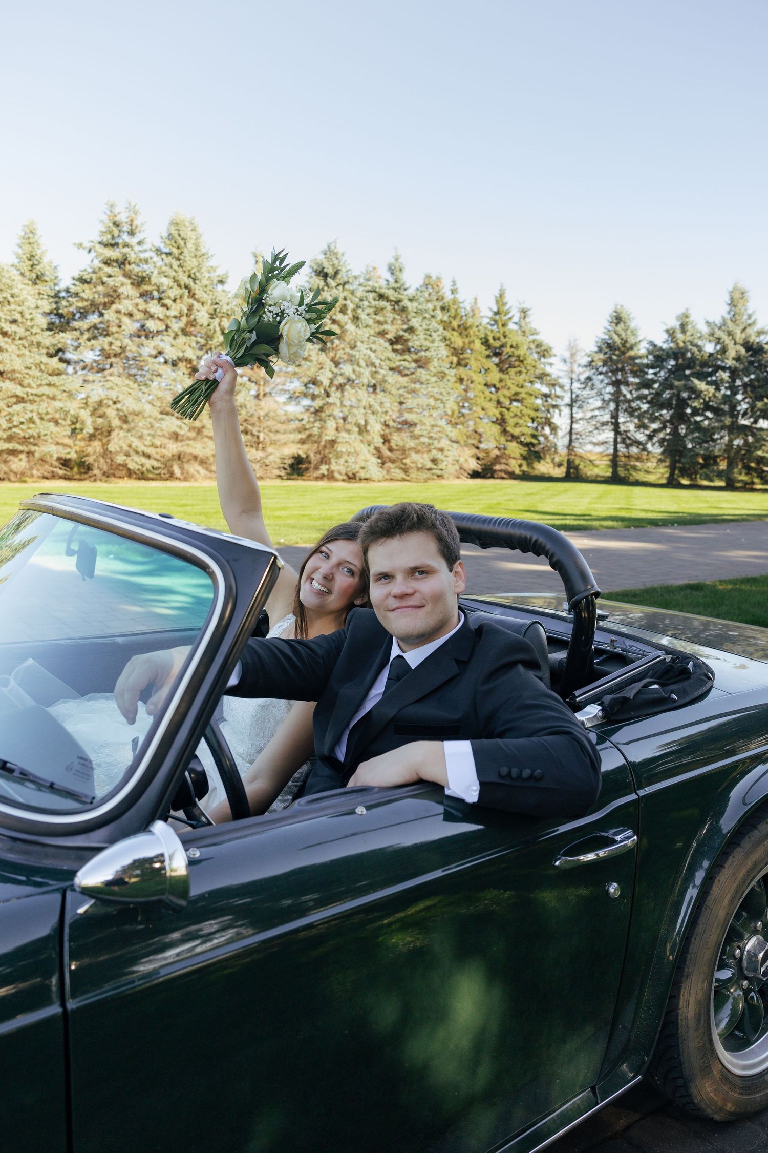 Happy newlywed couple in a dark green vintage convertible, bride holding bouquet aloft, groom smiling at camera.