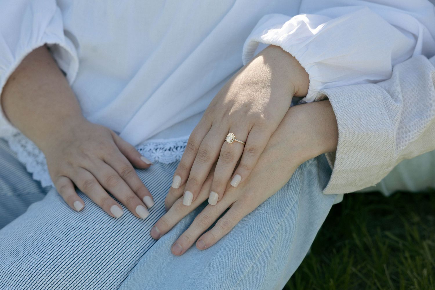 Two hands with white manicured nails, one wearing a gold wedding band, resting together on a lap in white clothing.