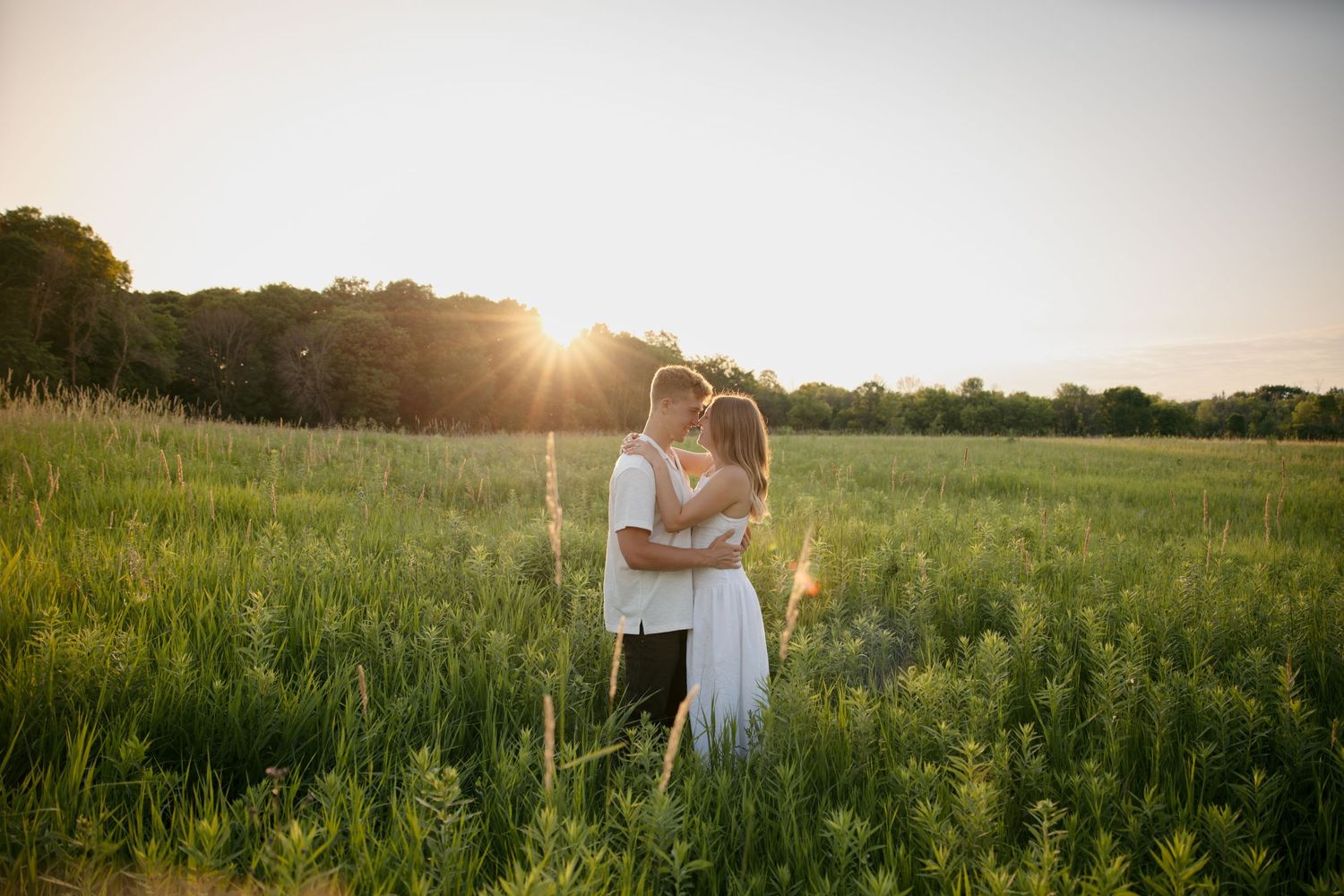Couple embracing in a lush green field at golden sunset, sun rays glowing behind them.
