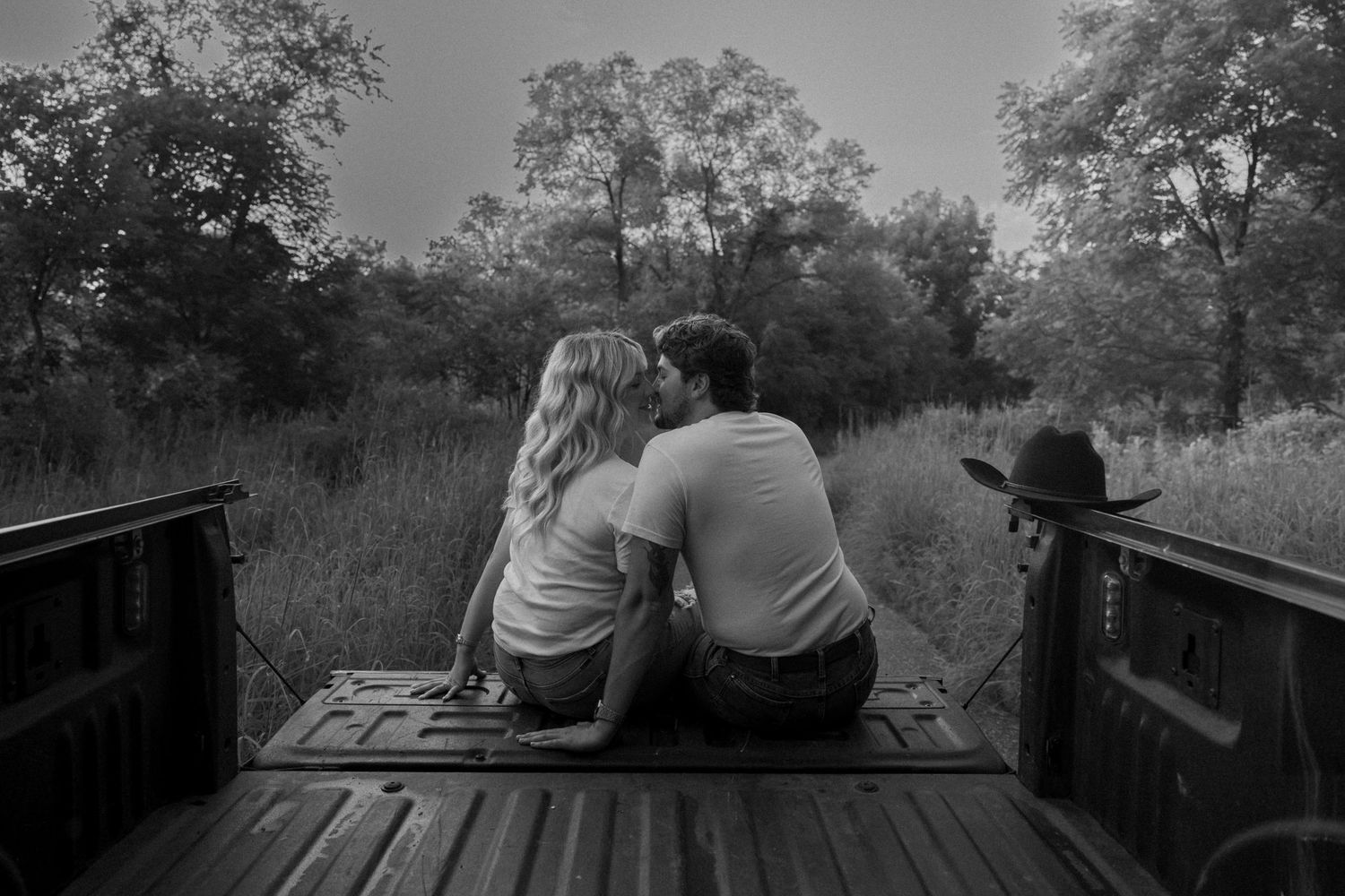 Black and white photo of a couple sitting closely together on a truck bed, surrounded by lush trees and tall grass.