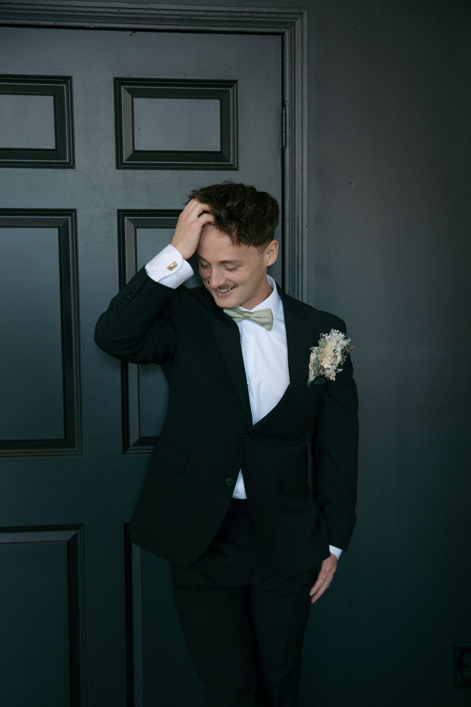 Groom in dark suit and bow tie laughing while leaning against dark door.