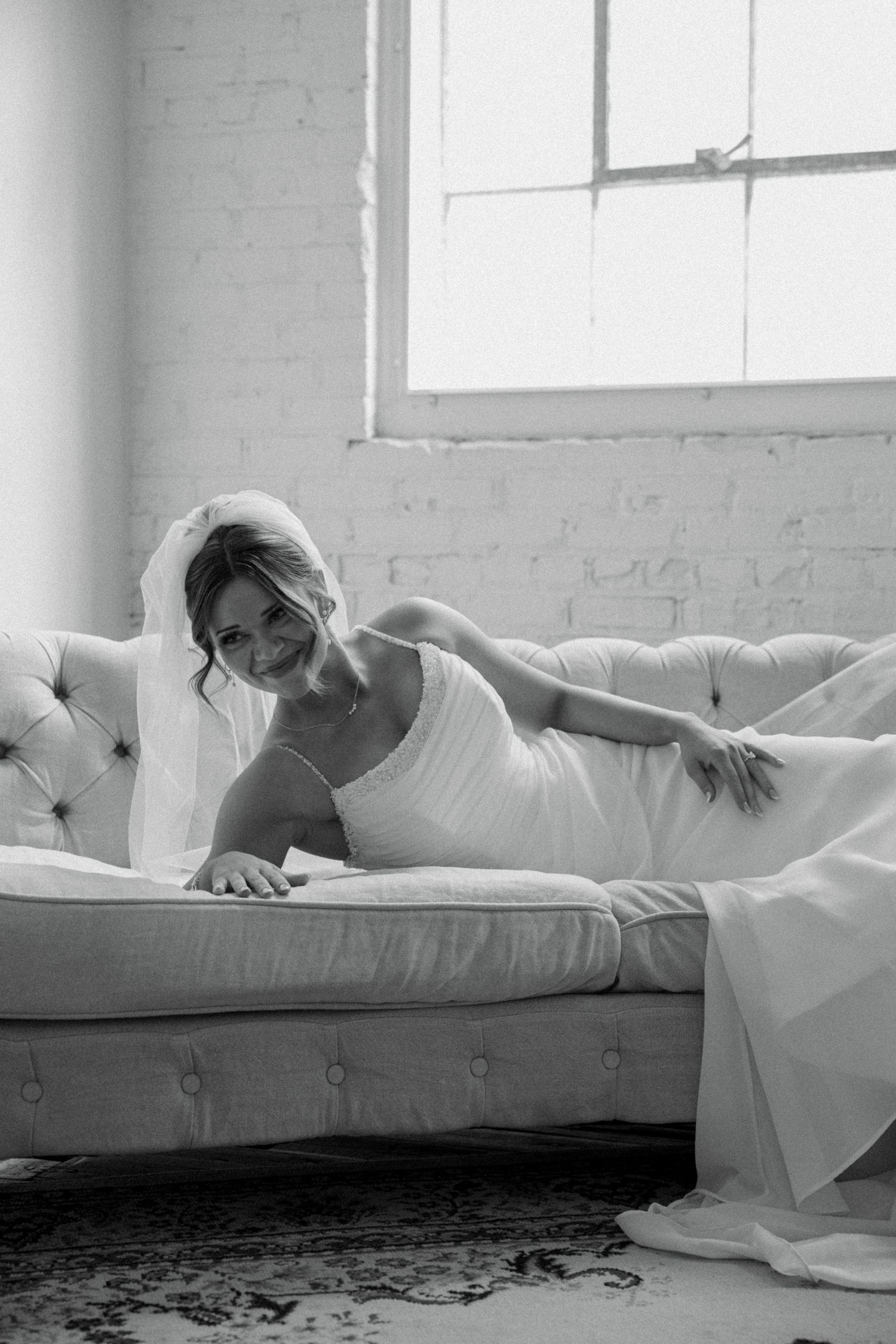 Bride in white gown and veil reclines gracefully on a tufted sofa in a bright, airy room in this black and white photo.