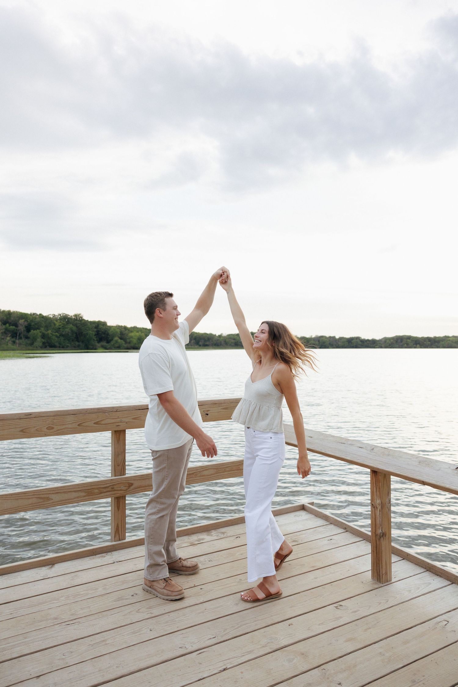 A couple dances joyfully on a wooden lakeside dock, surrounded by calm water and lush green trees under a cloudy sky.