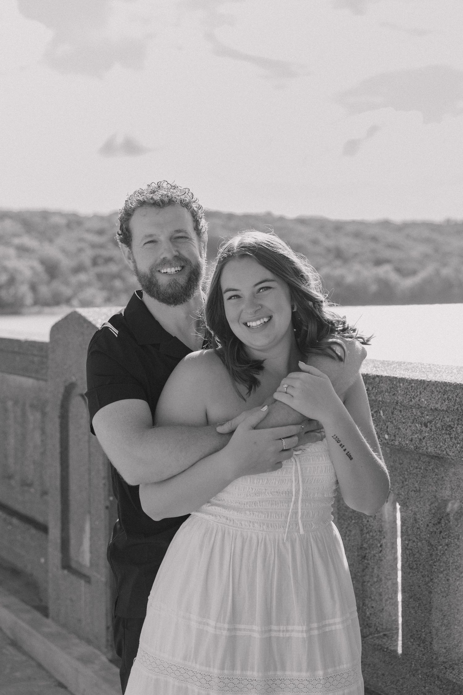 Black and white portrait of a happy couple hugging outdoors, woman in white dress smiling, man with beard behind her.