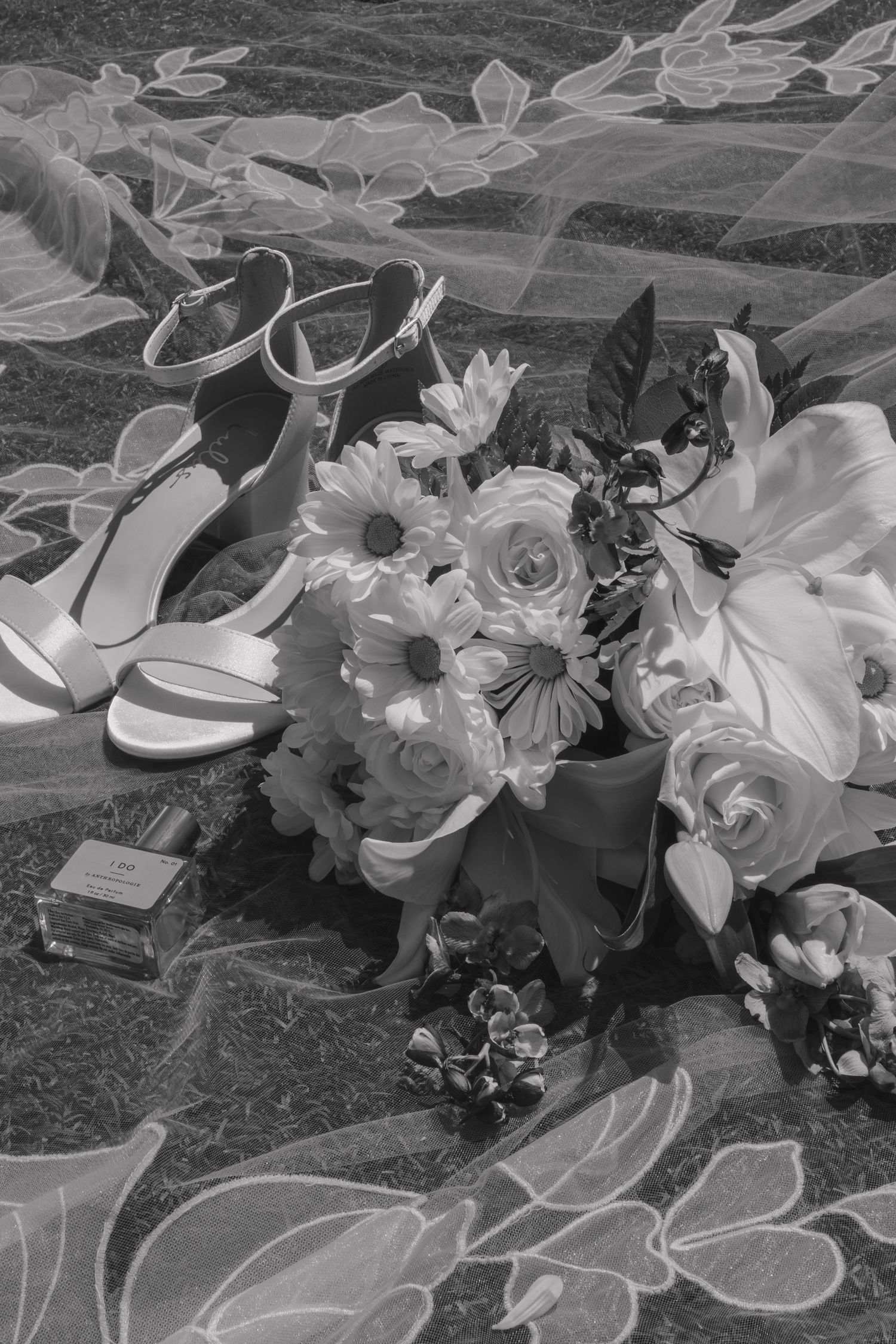 Black and white flat lay of bridal bouquet with roses, daisies, heels, and perfume on floral fabric.