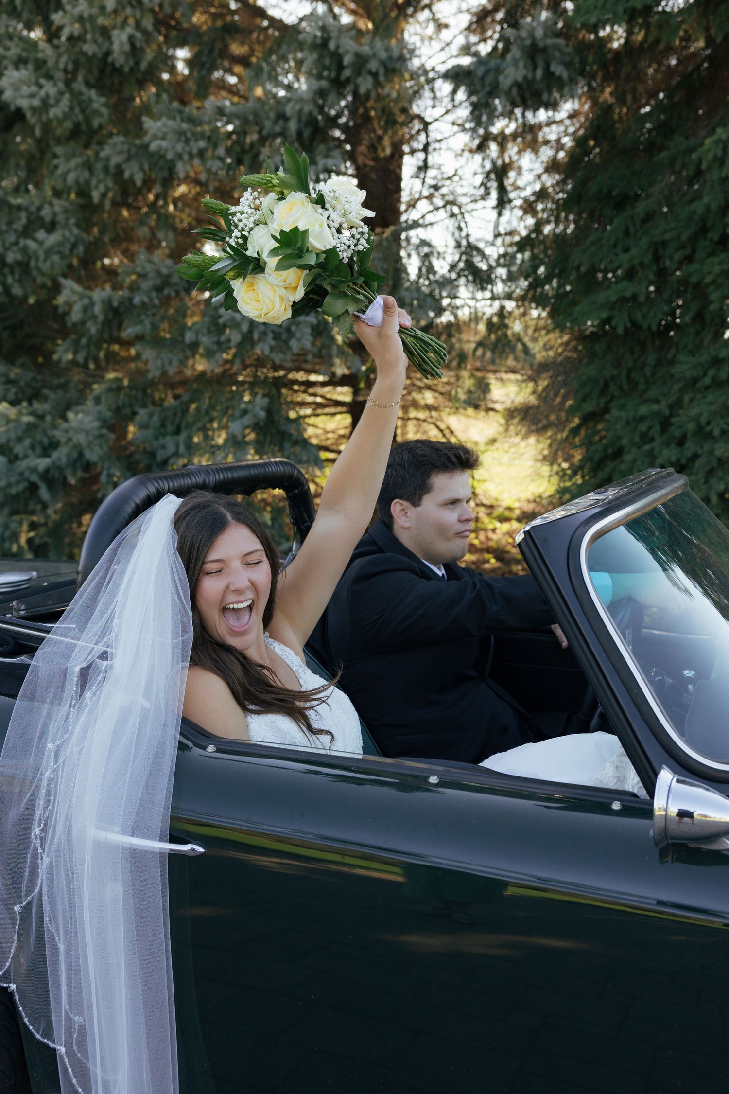 Joyful bride raising bouquet of white flowers while sitting in a vintage convertible car with groom on wedding day.