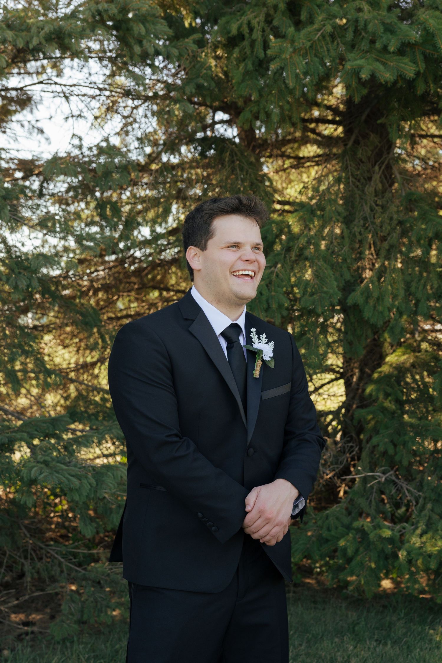Smiling groom in black suit with boutonniere stands outdoors before evergreen trees on his wedding day.