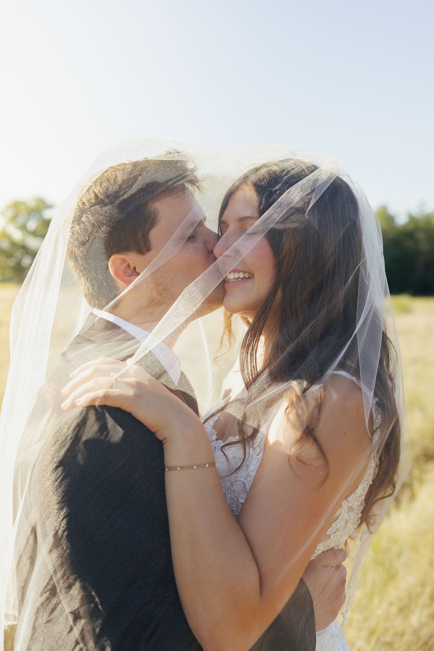 A bride and groom share a romantic kiss under a flowing veil in a sunlit field on their wedding day.