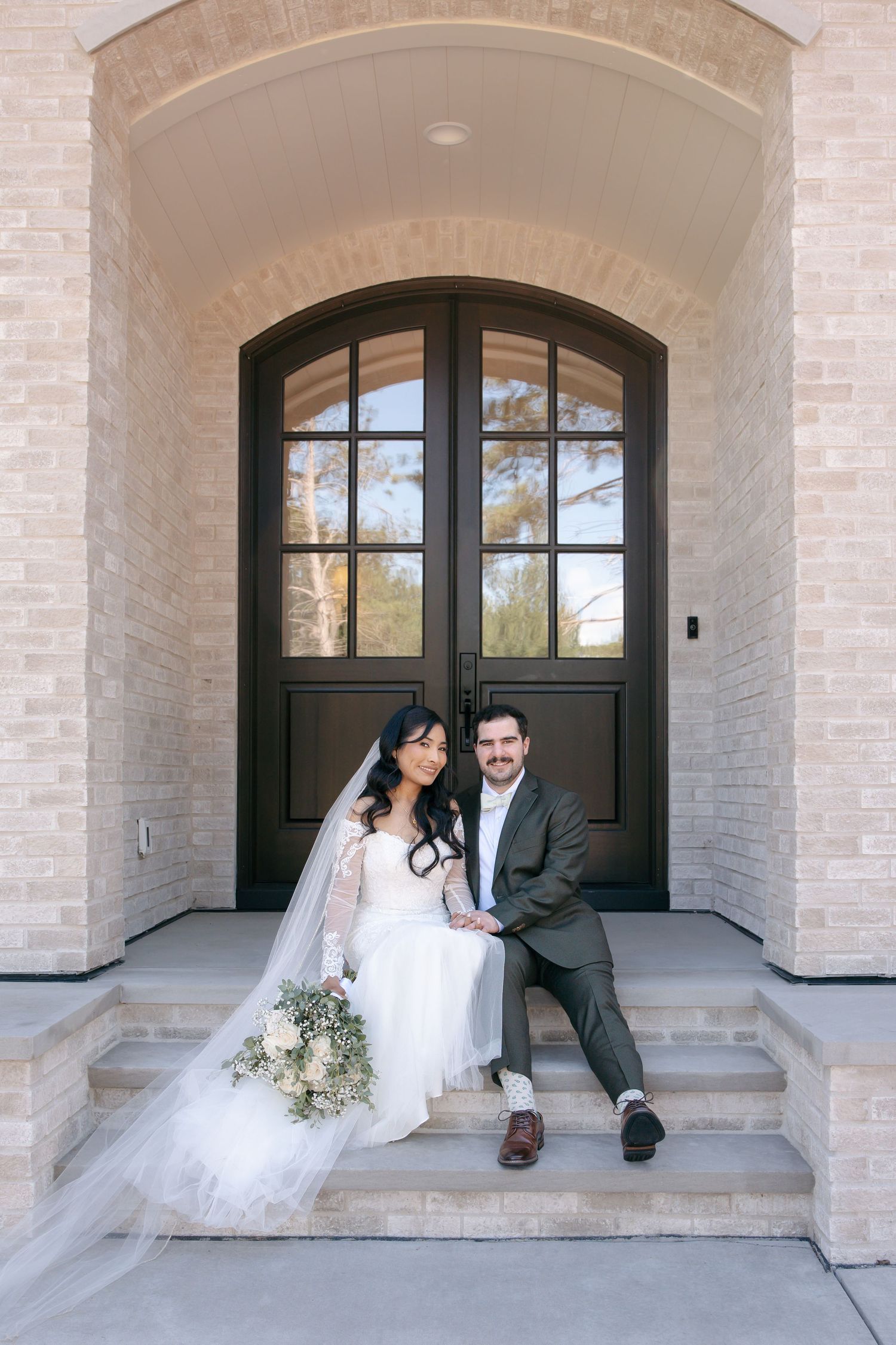 Bride and groom sitting on steps before elegant arched double doors of a brick building on their wedding day.