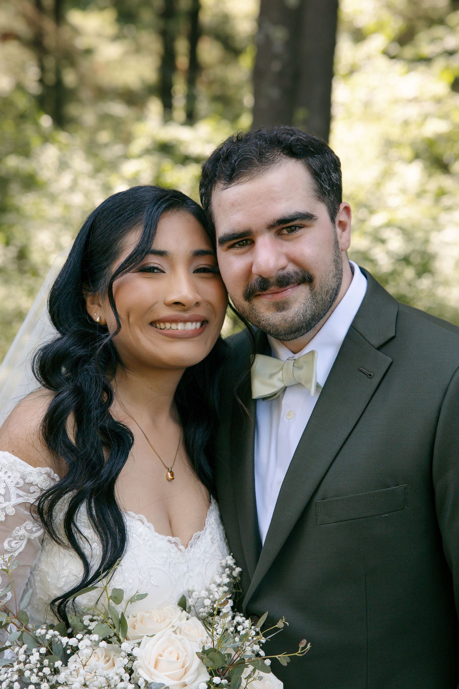 Smiling bride and groom pose together in forest, bride holding white rose bouquet.