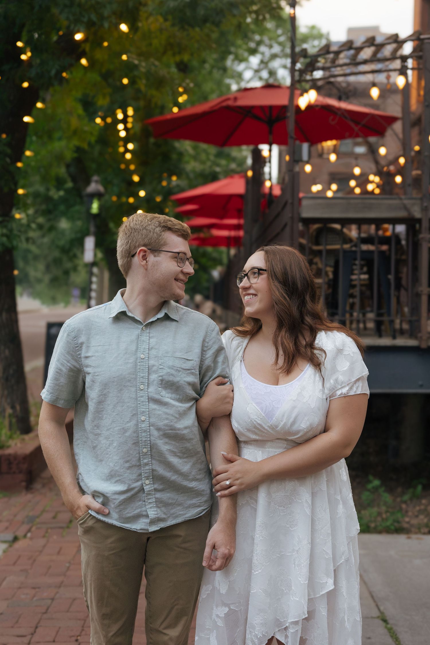 A smiling couple walks arm-in-arm on a brick path, with string lights and red patio umbrellas in the background.