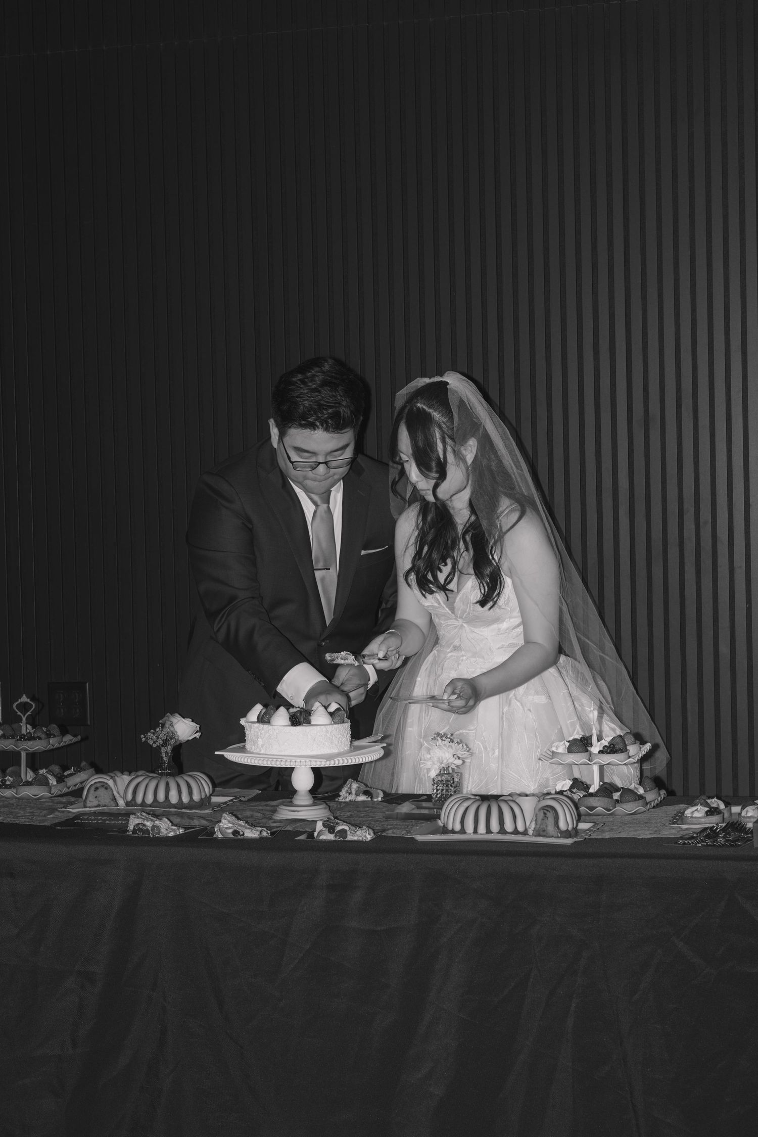 Black and white photo of a bride and groom cutting their wedding cake at the reception table.