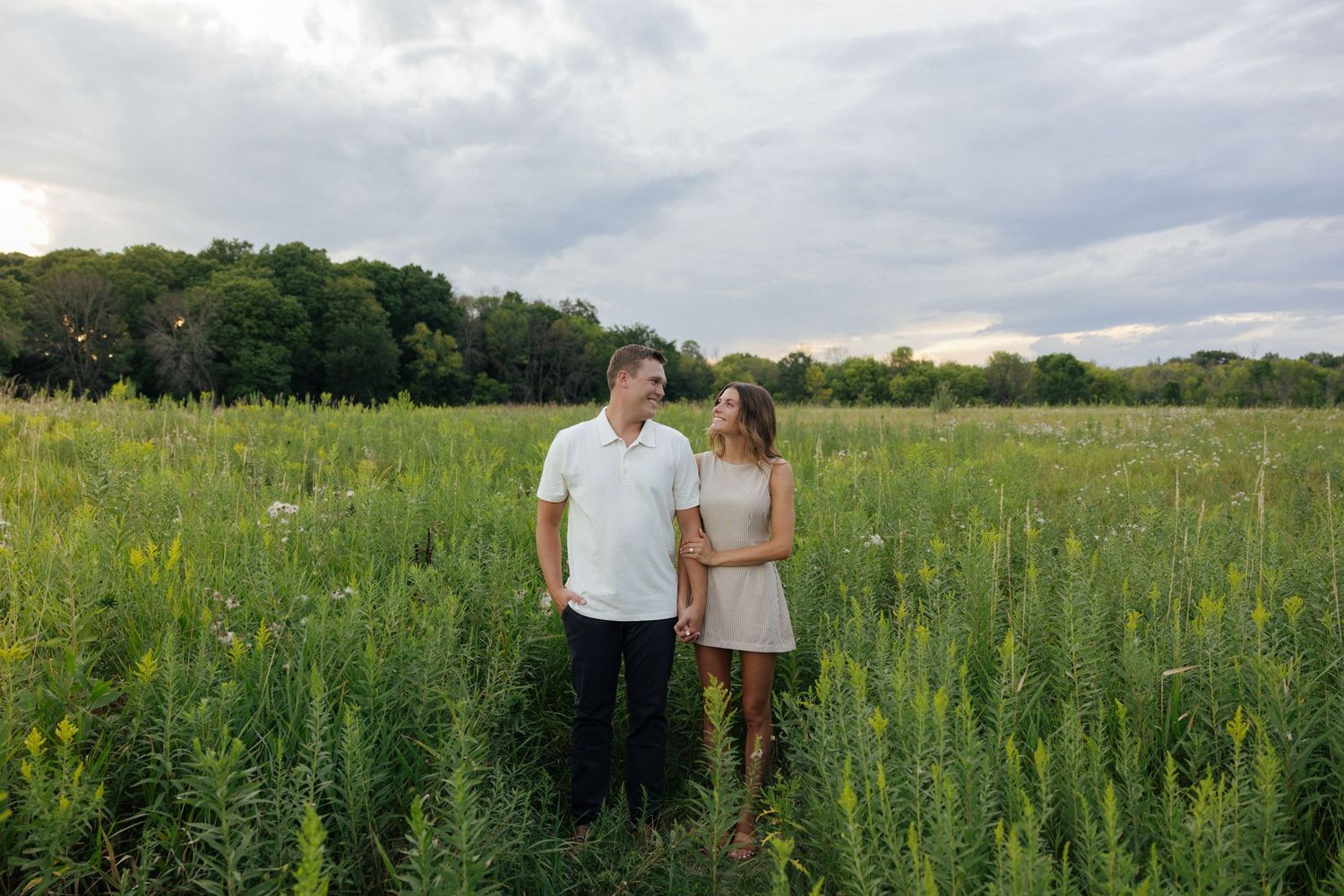 Couple standing together in a lush green meadow with wildflowers under a cloudy sky at sunset.