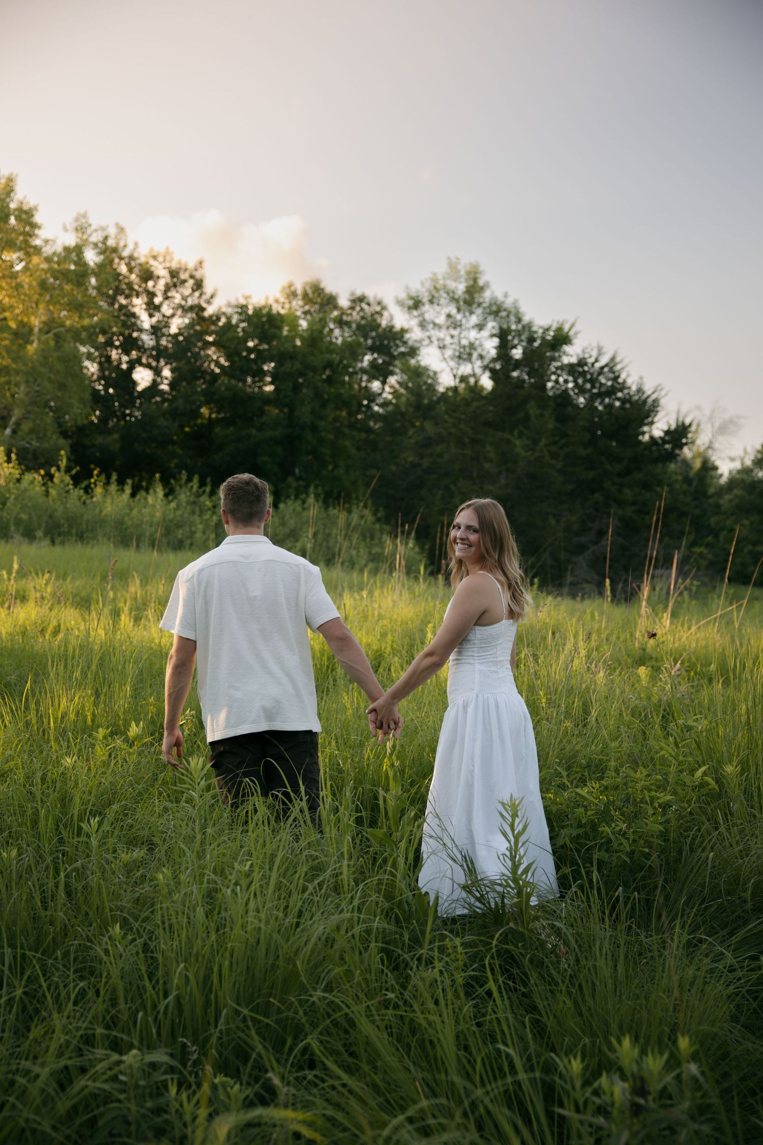 Couple holding hands walking through lush green field at golden hour, woman in white dress smiling back at camera.