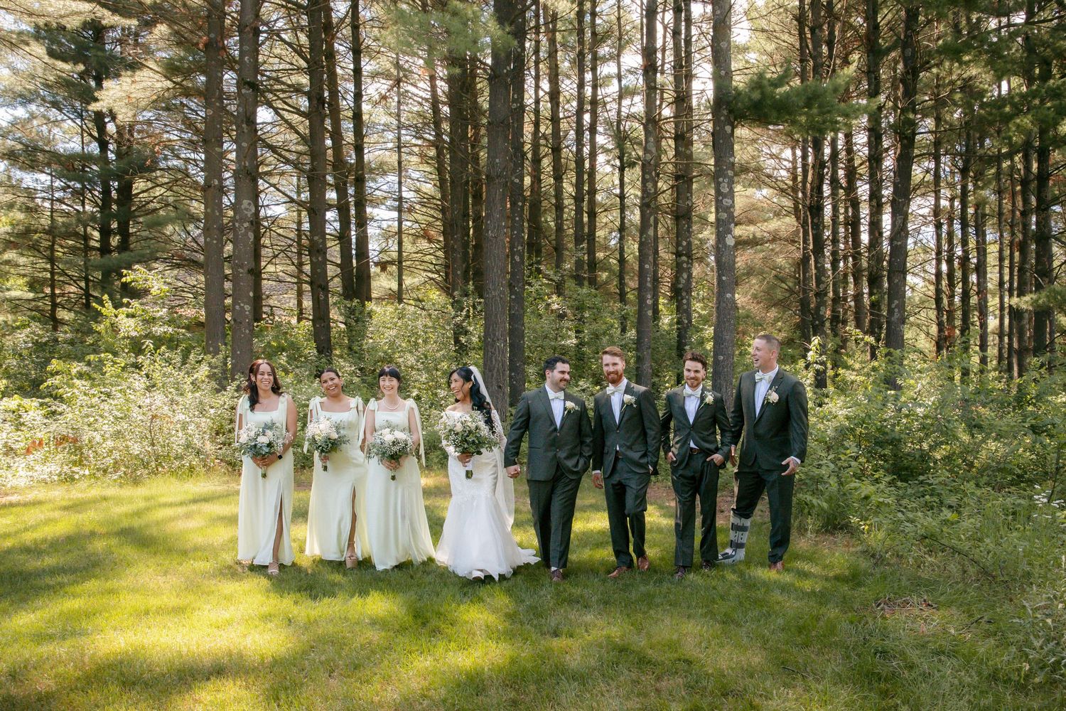 Wedding party in white and gray attire posed together in lush green forest setting.