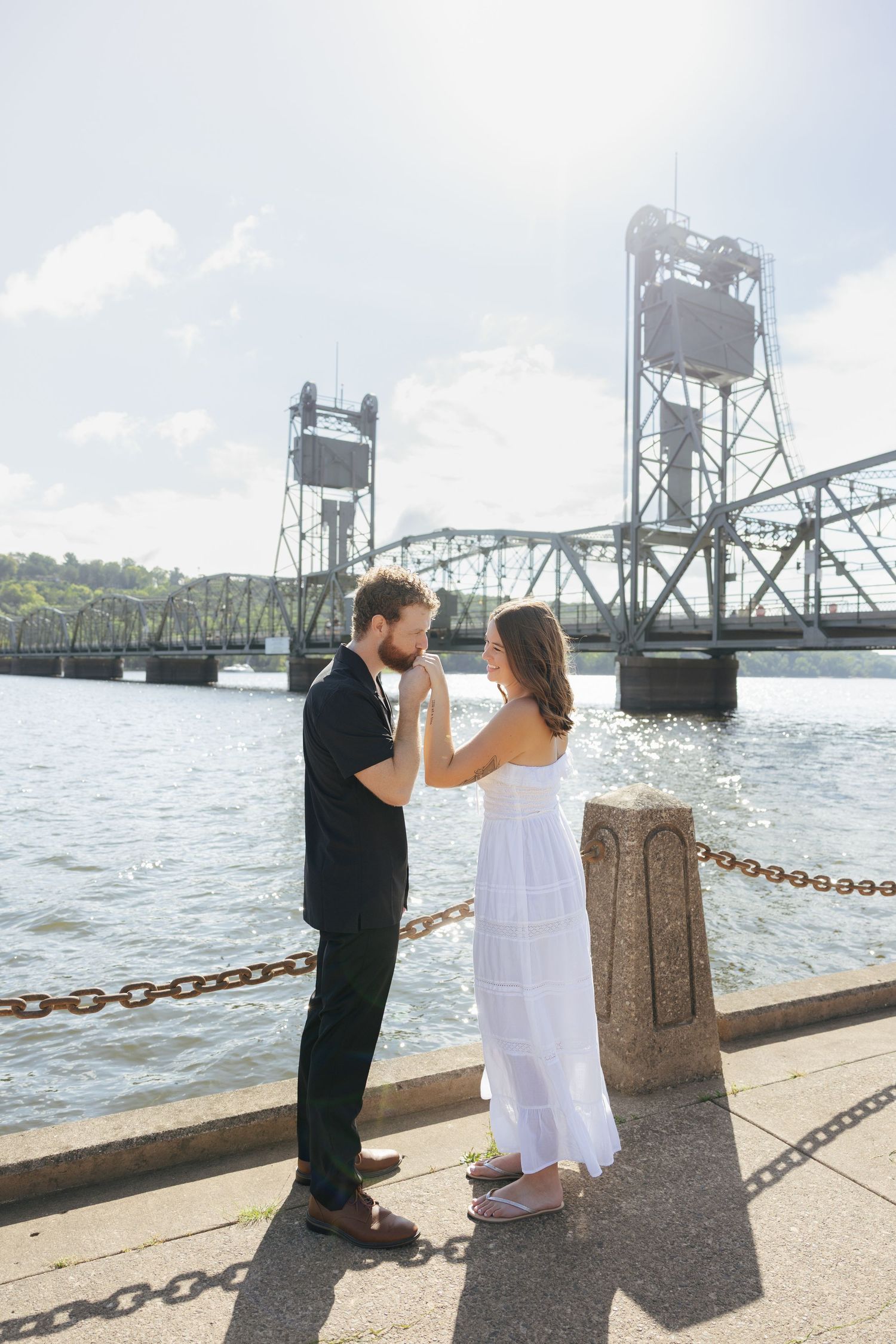 Couple holding hands by a river with a steel lift bridge backdrop on a sunny day.