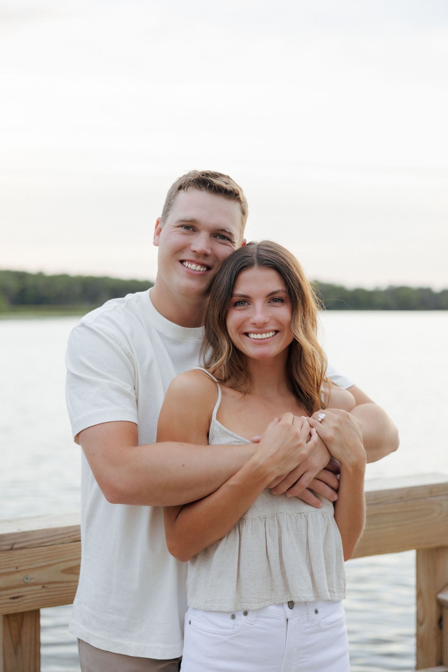 Smiling couple embracing on a wooden dock with a serene lake and trees in the background.