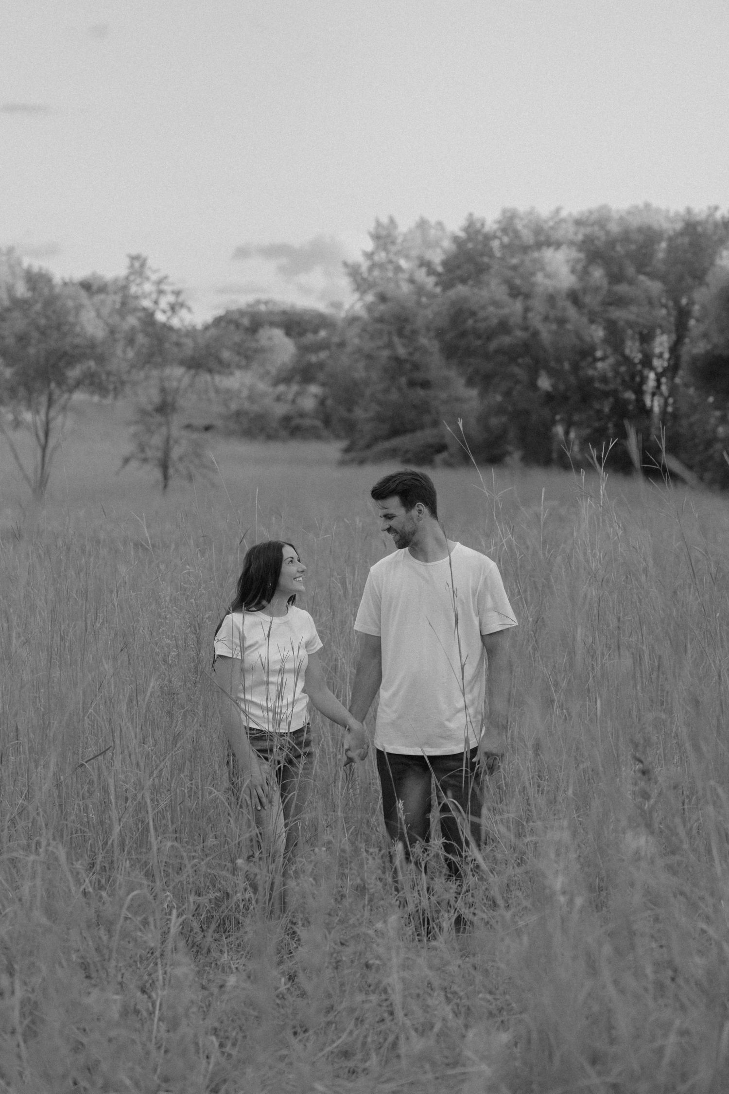 Black and white photo of a couple holding hands while walking through tall grass in a scenic field.