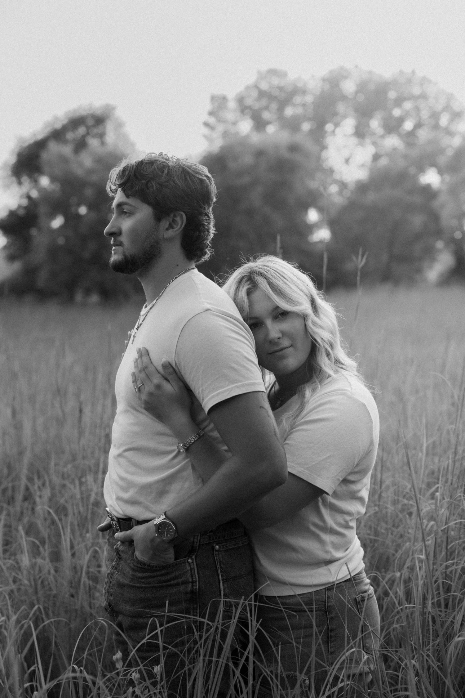 Black and white photo of a couple embracing in a field, woman resting her head on mans back.