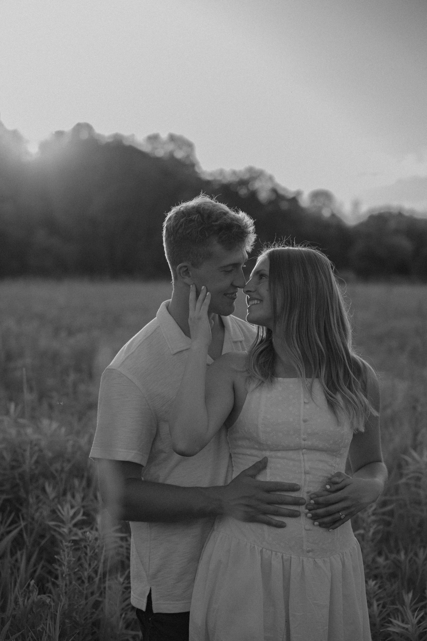 Black and white photo of a romantic couple nearly kissing in a sunlit field at dusk.